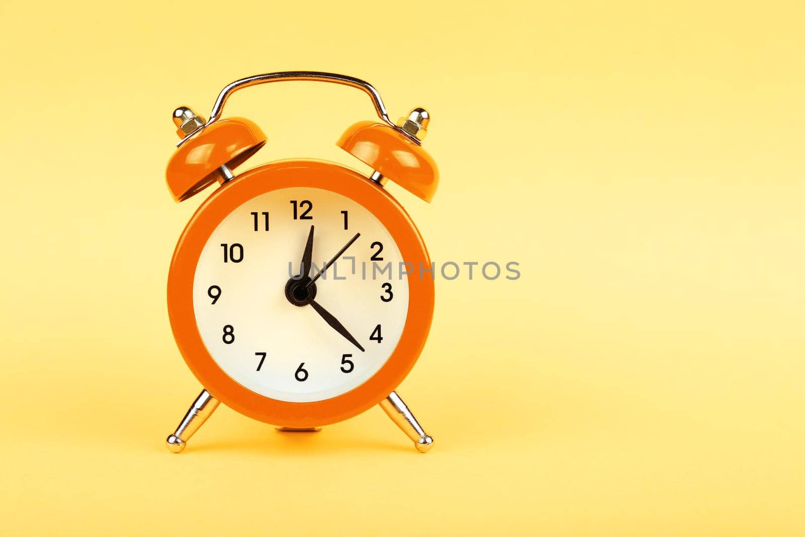 Close up one small orange metal twin bell retro alarm clock over pastel yellow paper background with copy space, low angle front view