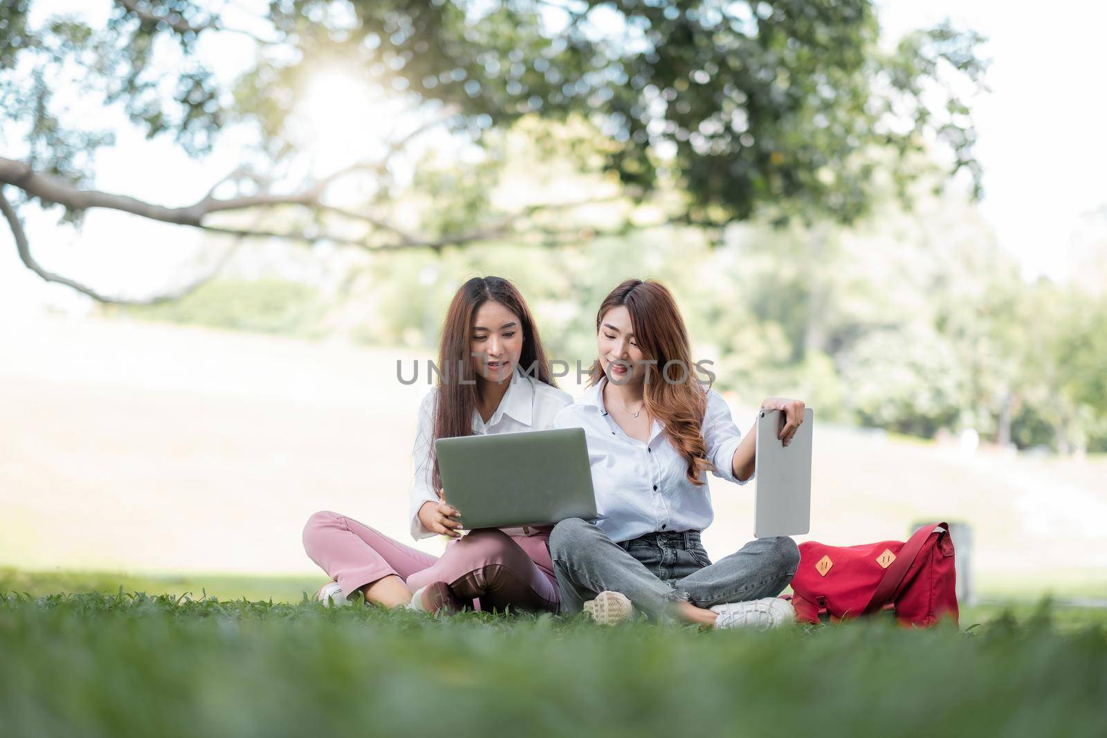Happy asian university sitting outside on campus. two girl working and studying together with laptop and digital tablet in the park by nateemee
