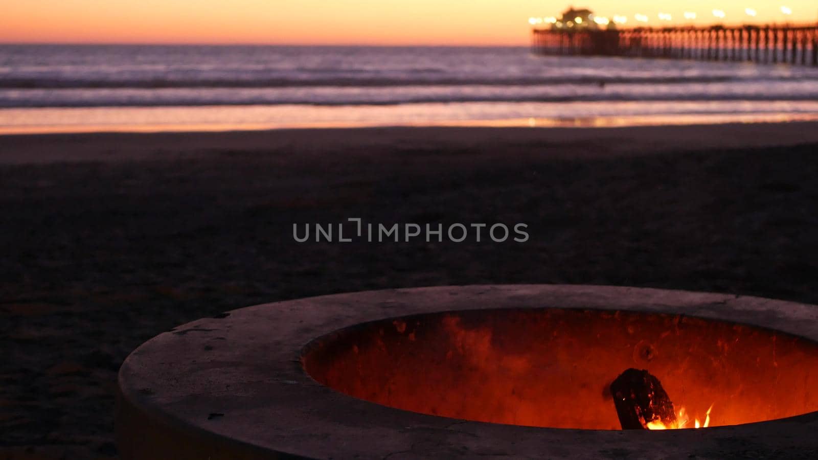 Campfire pit by Oceanside pier, California USA. Camp fire burning on ocean beach, bonfire flame in cement ring place for bbq, sea water waves. Romantic evening twilight sky, dusk after summer sunset.