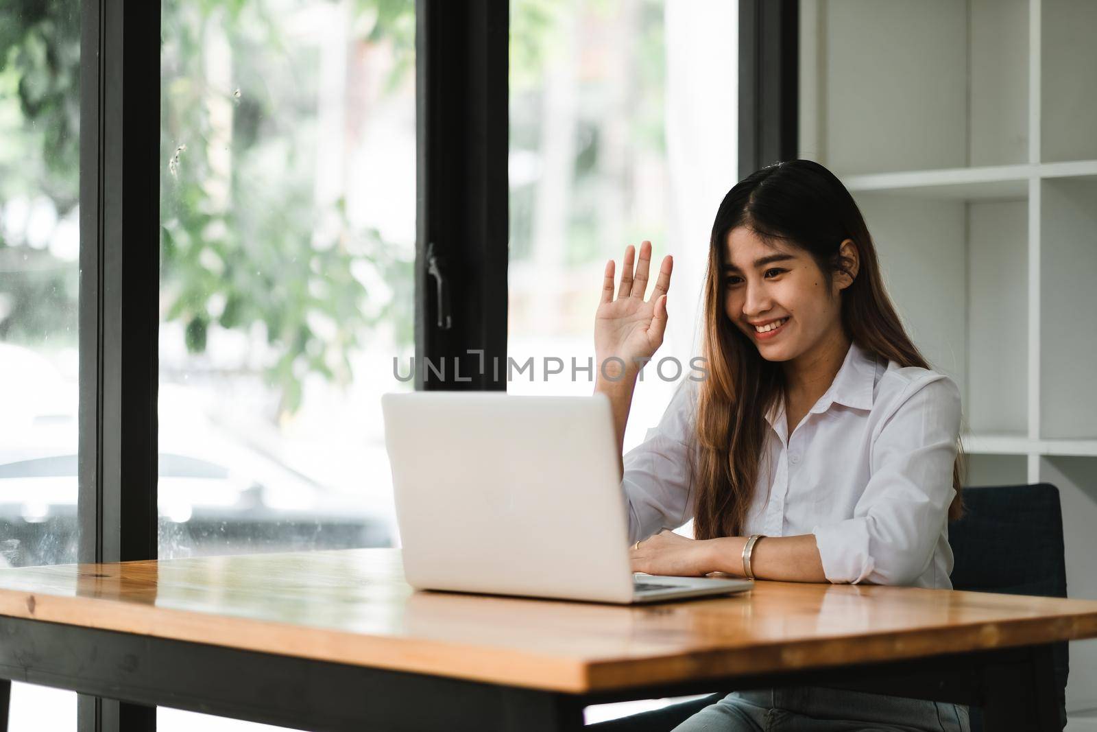 Smiling asian girl talking and waving hand hello during online video call via laptop computer, sitting in coffee shop. Cheerful business woman having conference in internet via portable notebook. by nateemee
