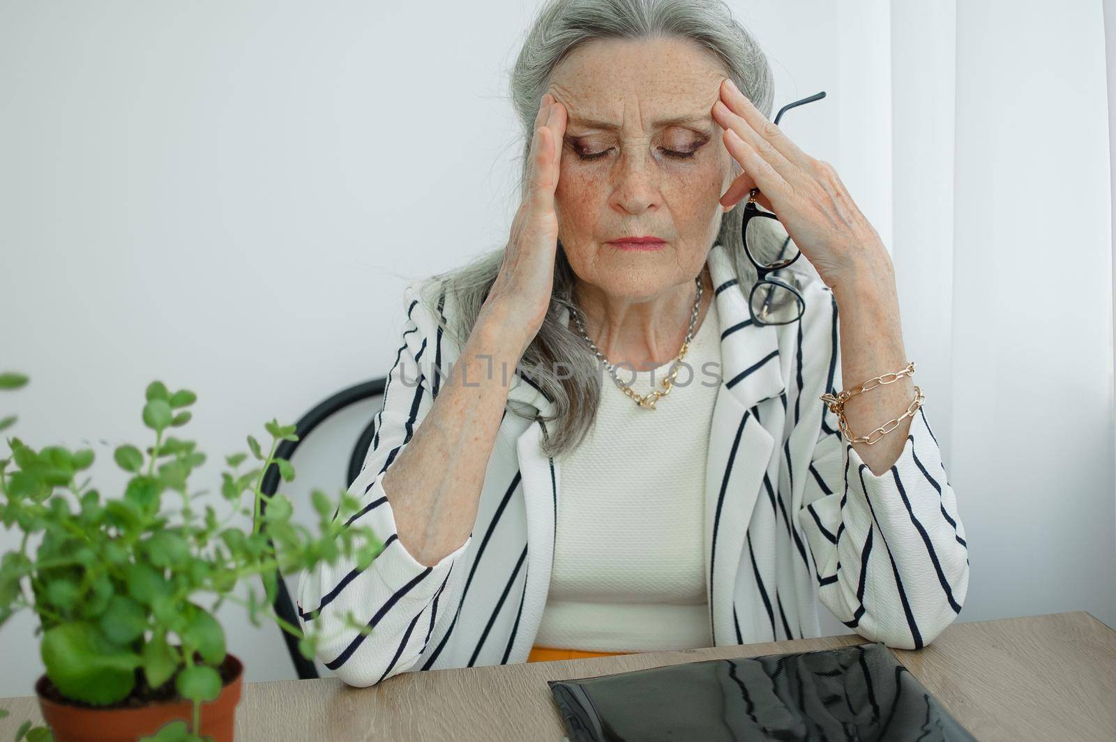 Tired senior grey haired businesswoman in striped jacket with eyeglasses is working in her office sitting at the desk and feeling bad due to menopause, menopause relief concept.