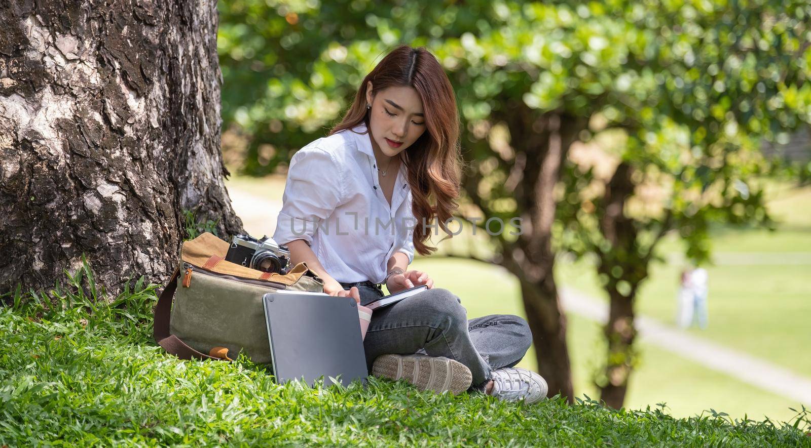 Image of joyful woman with diary book writing note while sitting on grass in park. by nateemee