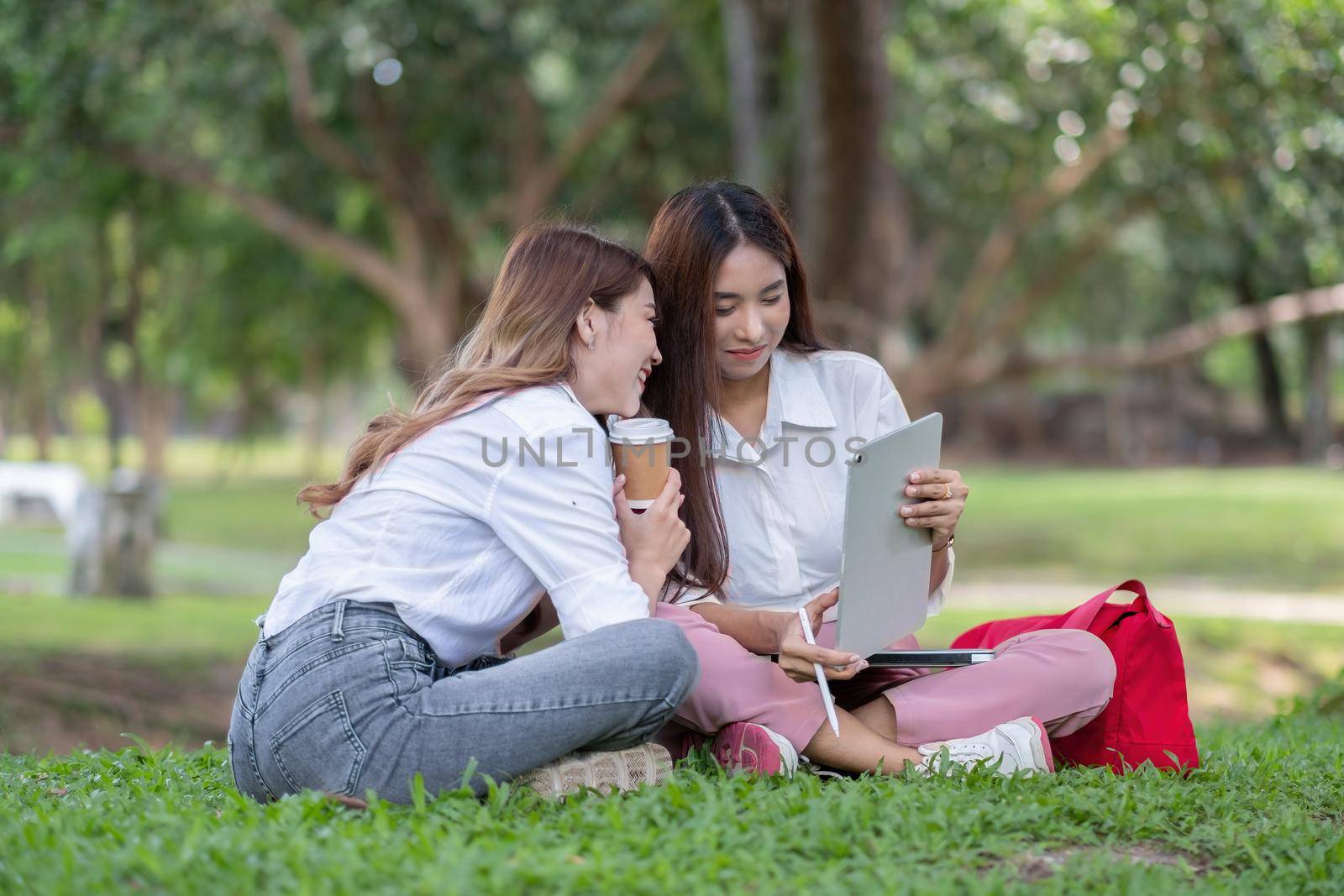 Close up happy asian university sitting outside on campus. two girl working and studying together with laptop and digital tablet in the park by nateemee