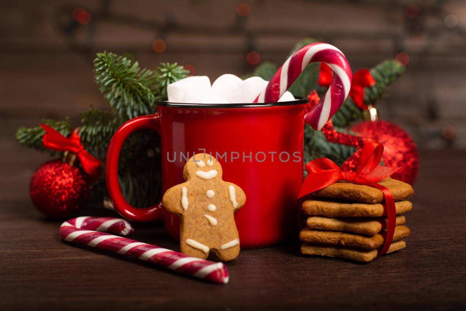 Cocoa in red mug with marshmallows candy cane fir tree branches and red baubles on dark wooden background with bokeh lights