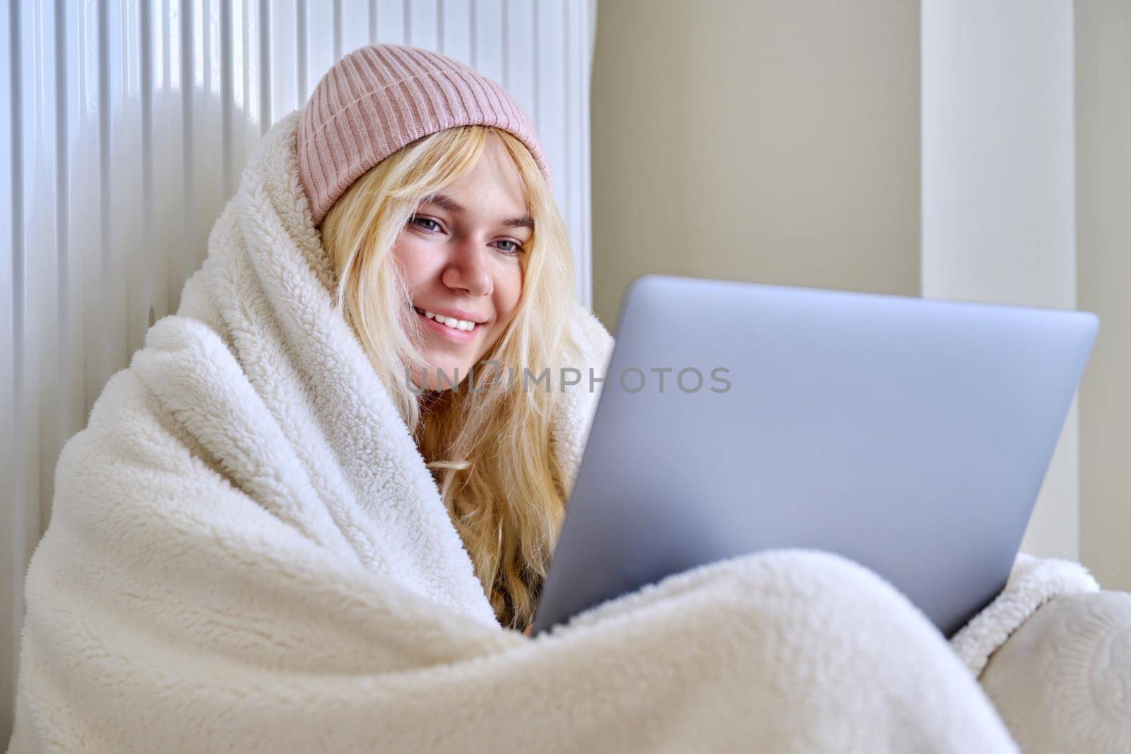 A young female teenager frozen in house in winter cold season, warming up with a warm blanket, hat, central heating radiator, looking at laptop screen, student, having rest