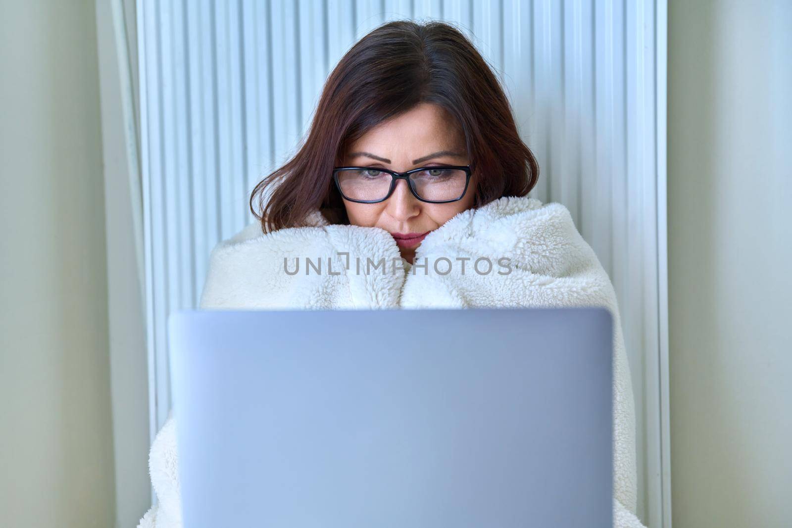 A frozen mature woman working at home with a laptop, warming herself with a blanket and heating radiator. Autumn winter cold season