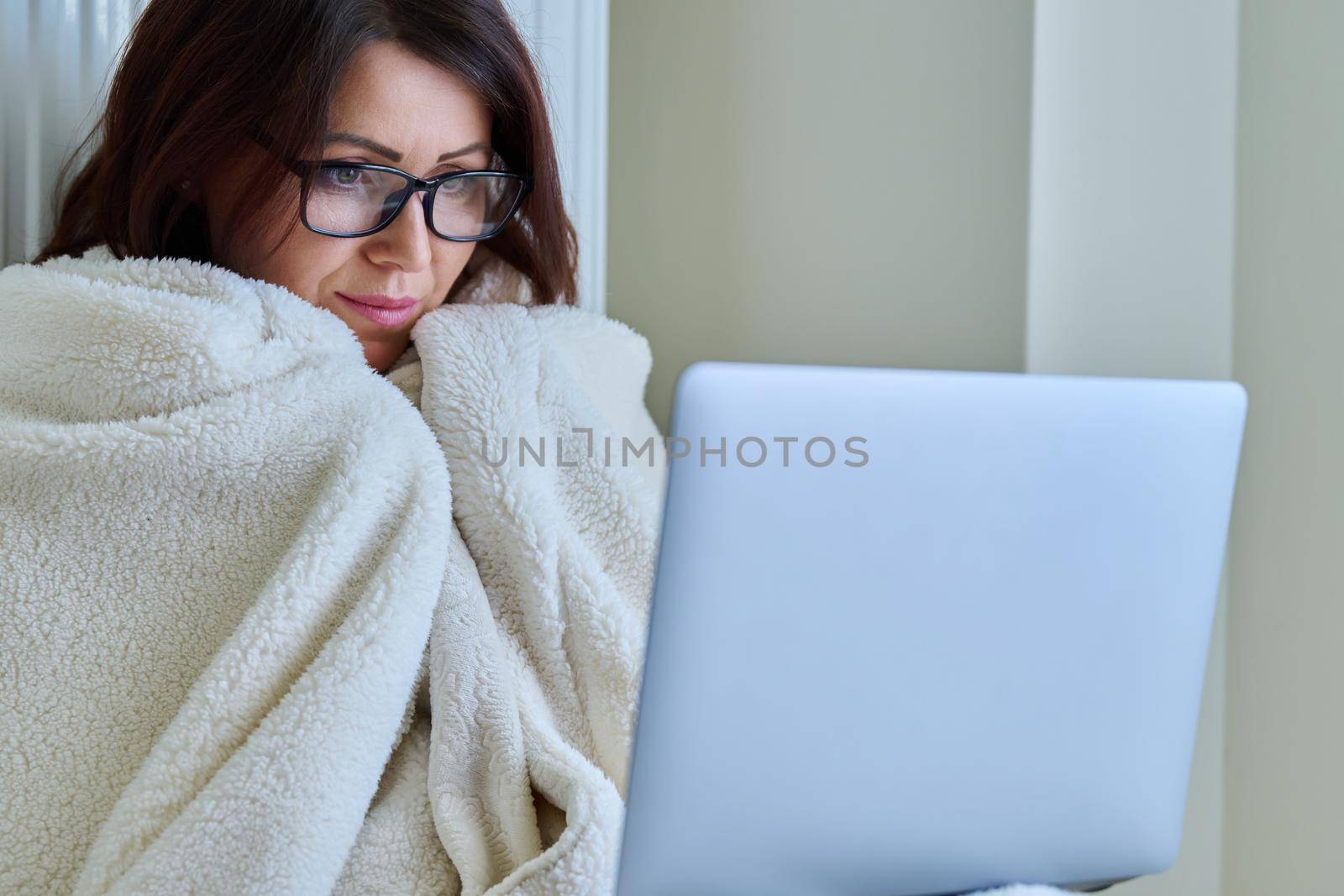 A frozen mature woman working at home with a laptop, warming herself with a blanket and heating radiator. Autumn winter cold season