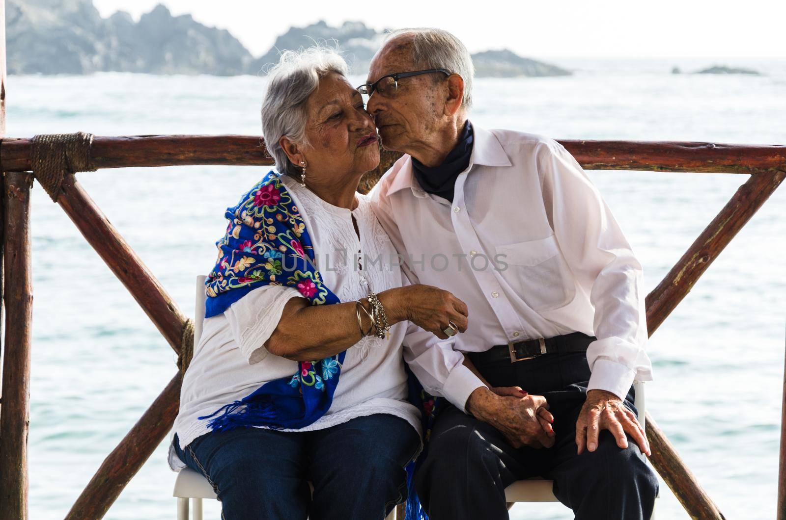 Elderly couple sitting on chairs kissing by Peruphotoart