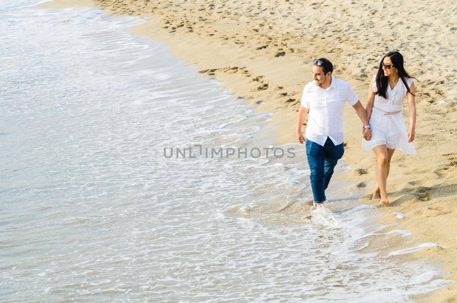 Barefoot young couple walking hand in hand along a beach at the edge of the surf. by Peruphotoart