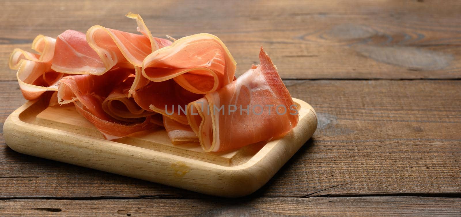 sliced thin slices of prosciutto on wooden brown board, wooden table background