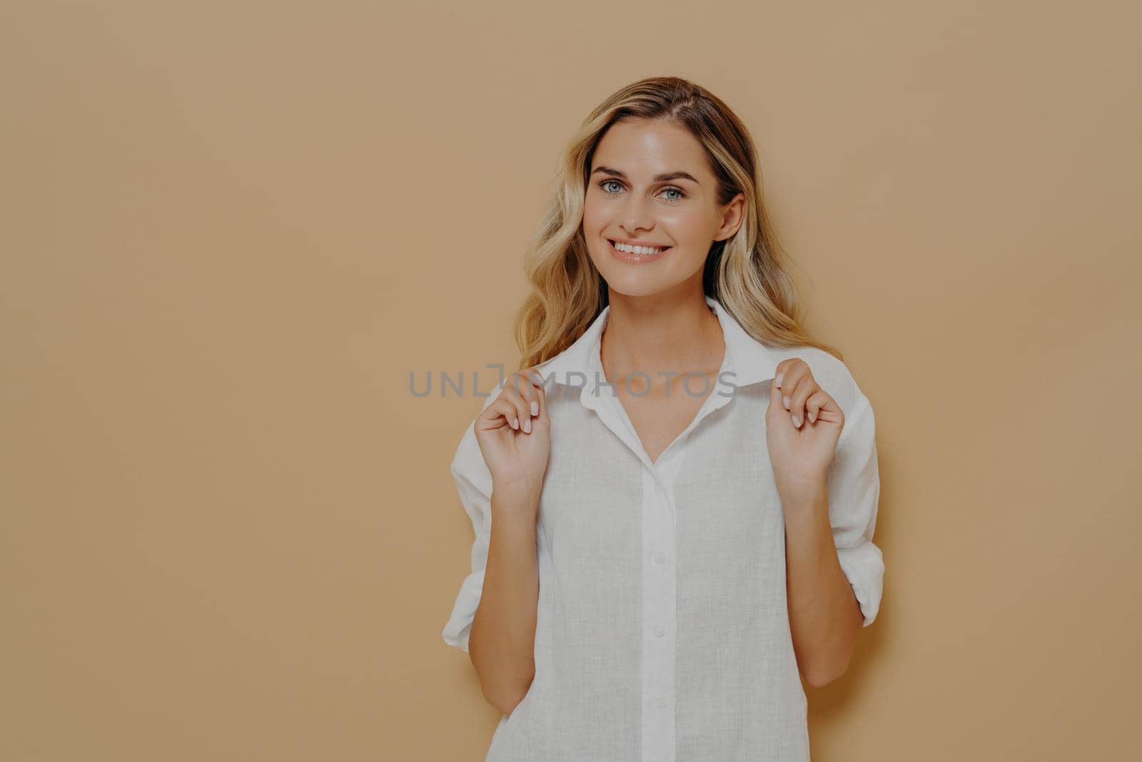 Expressing positivity. Young happy good looking woman with blond dyed hair in white shirt smiling cheerfully and looking at camera with playful facial expression,posing isolated over wall in studio