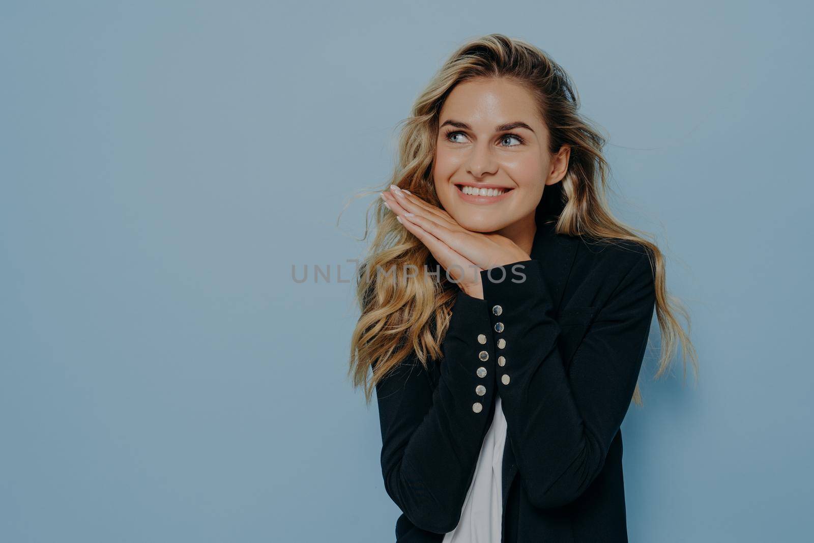 Delightful young european woman with blonde wavy hair wearing black blazer over white tshirt feeling happy of hearing compliment from friends, smiling and looking upside. Positive human emotions