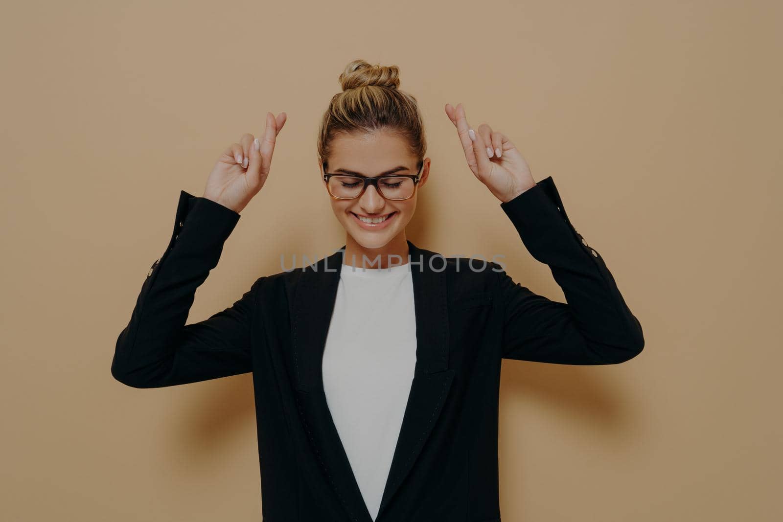 Hopeful young woman standing with hands up and crossed fingers in luck gesture, female in black stylish blazer with blonde hair tied in cute neat bun making wish with closed eyes. Body language