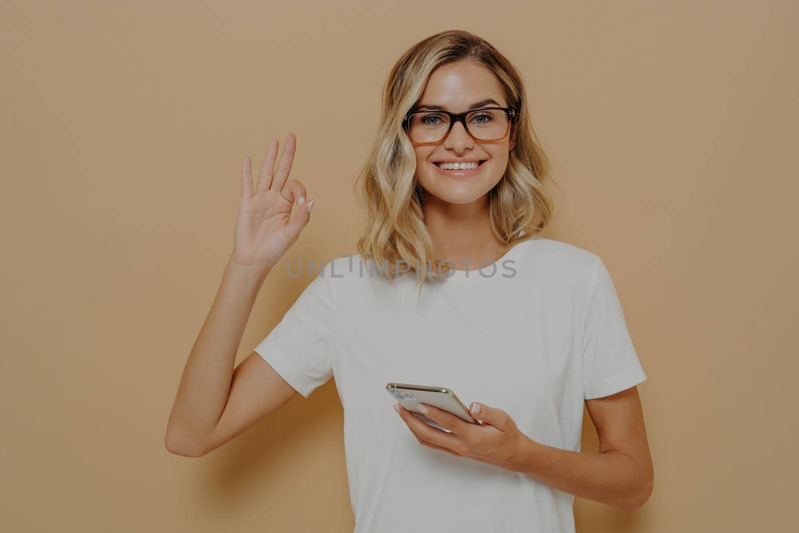 Everything ok. Young happy woman smiling at camera and showing okay gesture with modern smartphone in hand, dressed in white tshirt. Cheerful female in spectacles using cellphone and being sitisfied