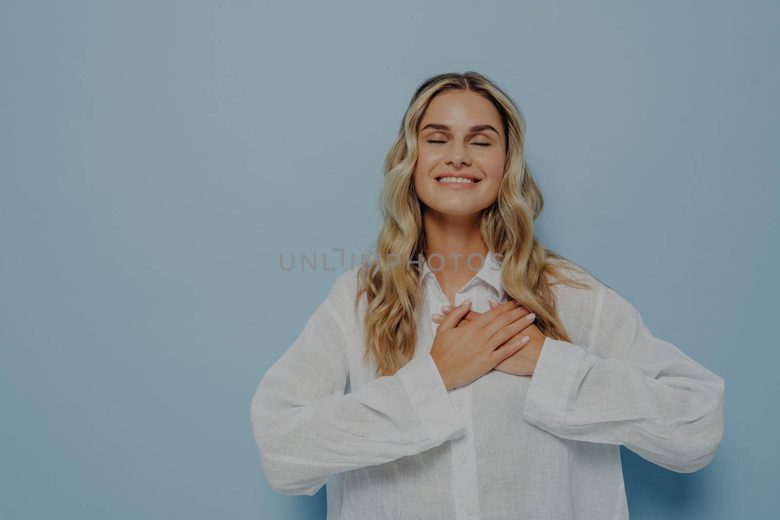 Pleased happy thankful woman with blond wavy hair in white oversize shirt gesturing appreciation and love and gratitude by holding hands together on chest, eyes closed, isolated over blue wall