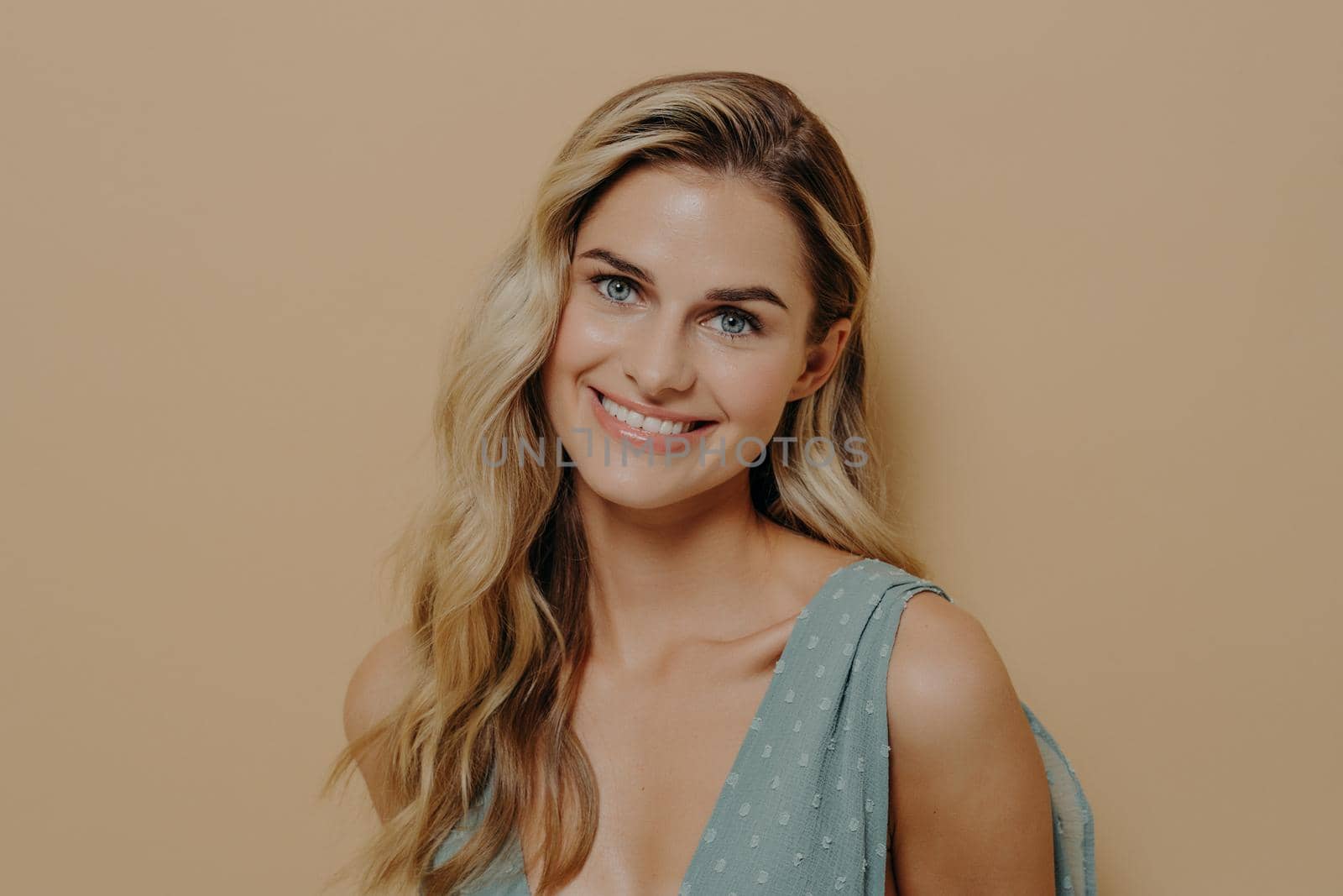 Beautiful tender young blond girl in blue dress with fresh radiant skin posing in front of the camera with beaming smile. Photo taken in studio on beige isolated background with copy space for text