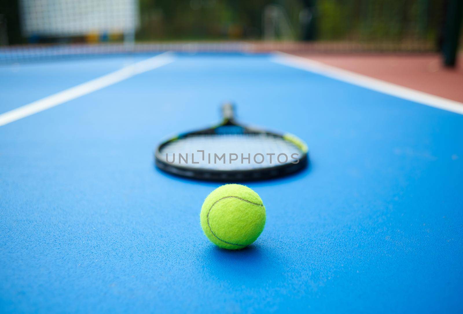 Frontview of yellow tennis ball is laying near professional racket on blue carpet of opened tennis cort. Contrast image with satureted colors and shadows. Concept of sport equipment photo.