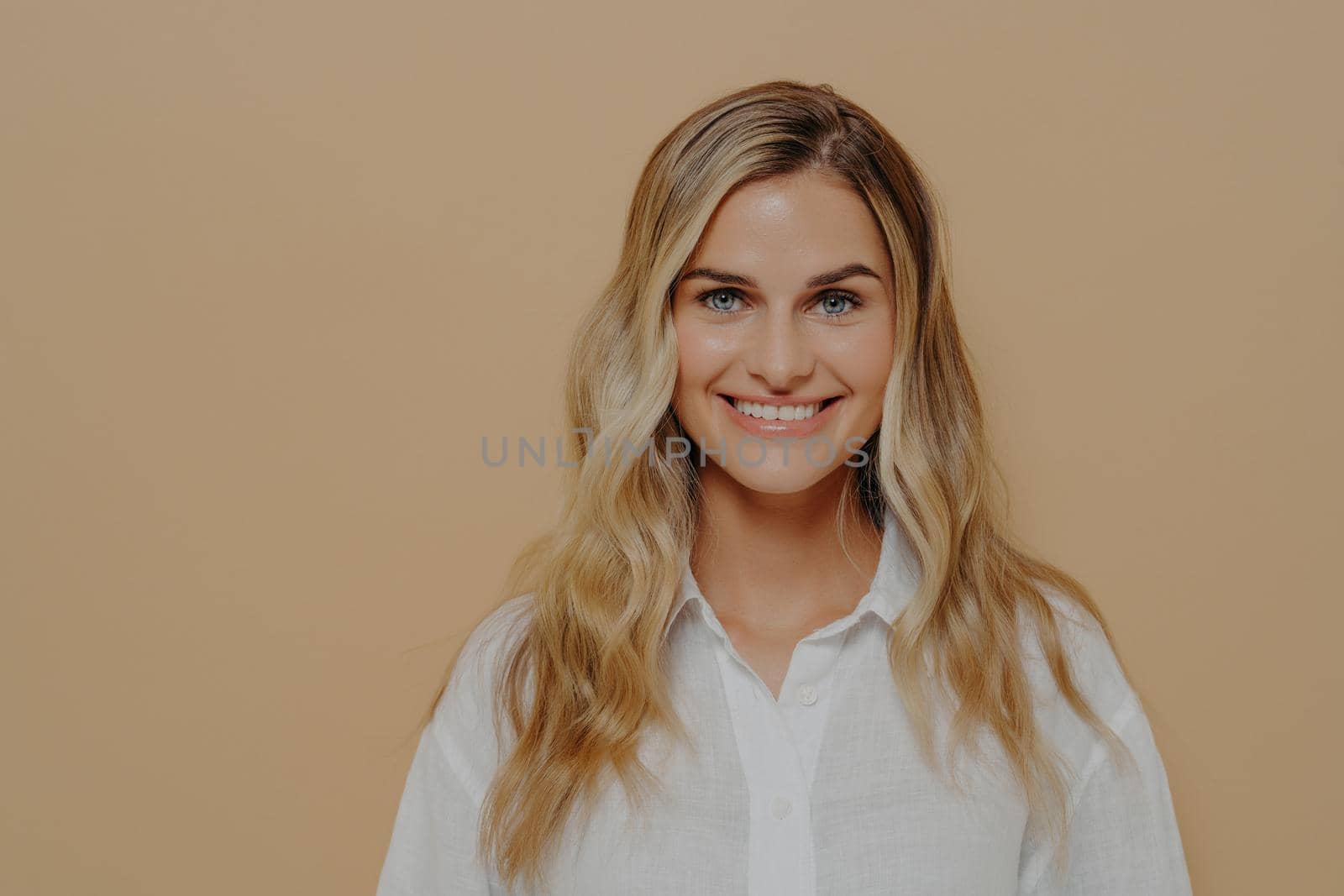 Portrait of calm blonde teenage girl in white shirt smiling and looking straight at camera, wide toothy smile, radiating happiness and joy while standing isolated next to orange background