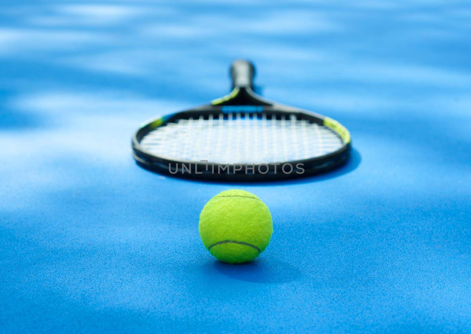 Yellow tennis ball is laying near professional racket on blue cort carpet. Made for playing tennis. Contrast image with satureted colors and shadows. Concept of sport equipment photo.