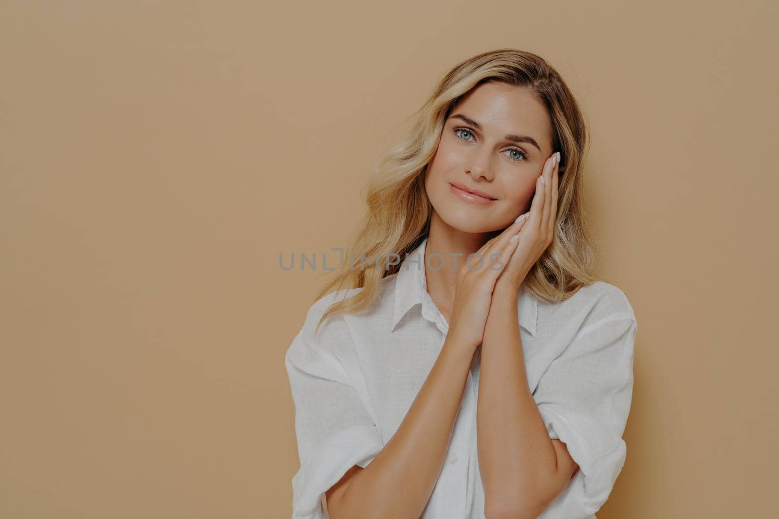 Romantic young blonde lady in casual white shirt keeping folded hands near her face and looking at camera with gentle smile, posing over beige background with copy space. Human face expressions