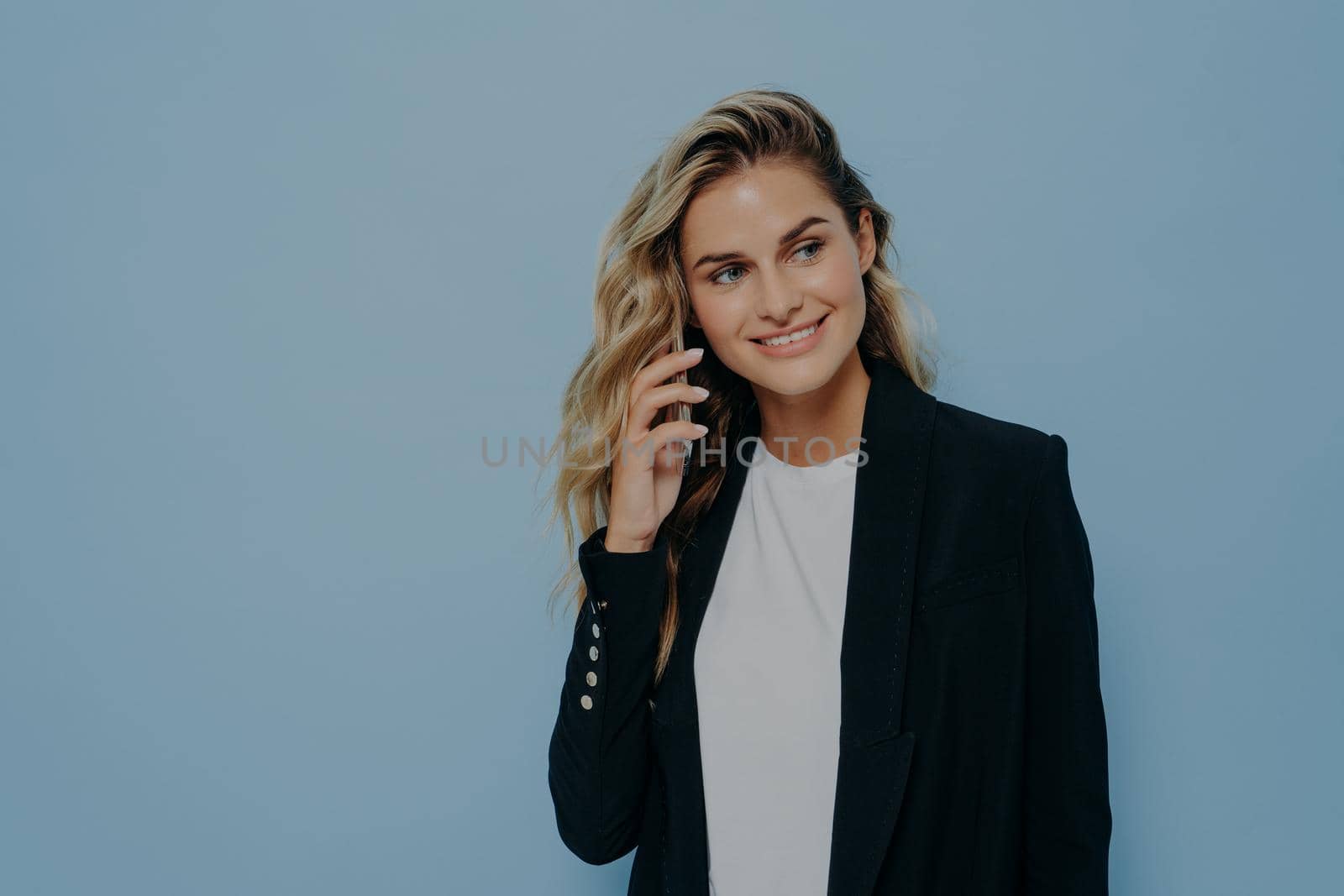 Blonde business woman in black jacket talking on mobile phone with family, happy to hear good news, listening and looking sideways while standing isolated on blue background. Communication concept