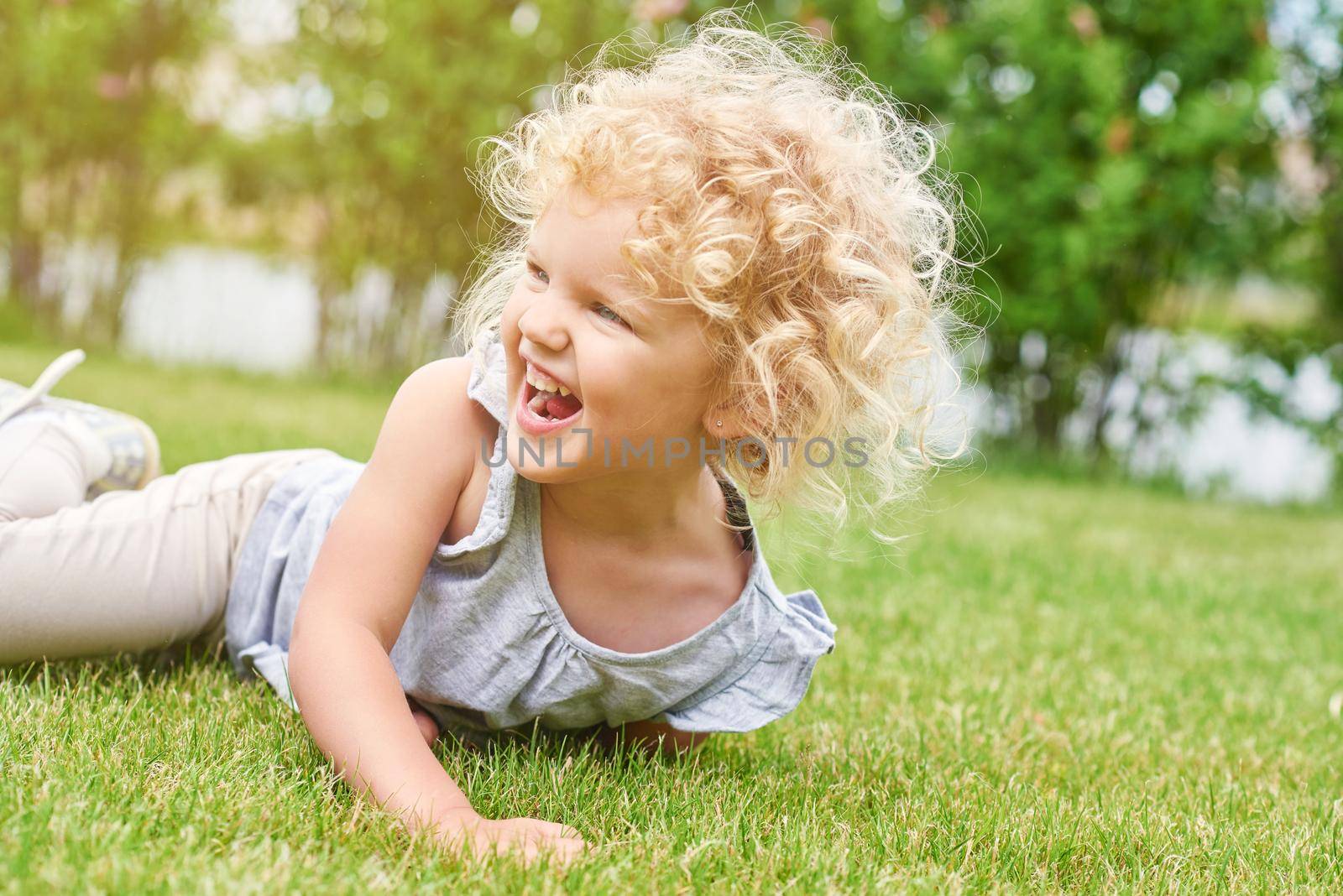 Shot of a cute little girl with blonde curly angelic hair lying on the grass laughing happily looking away copyspace nature enjoyment emotions kids children family recreation summer concept.