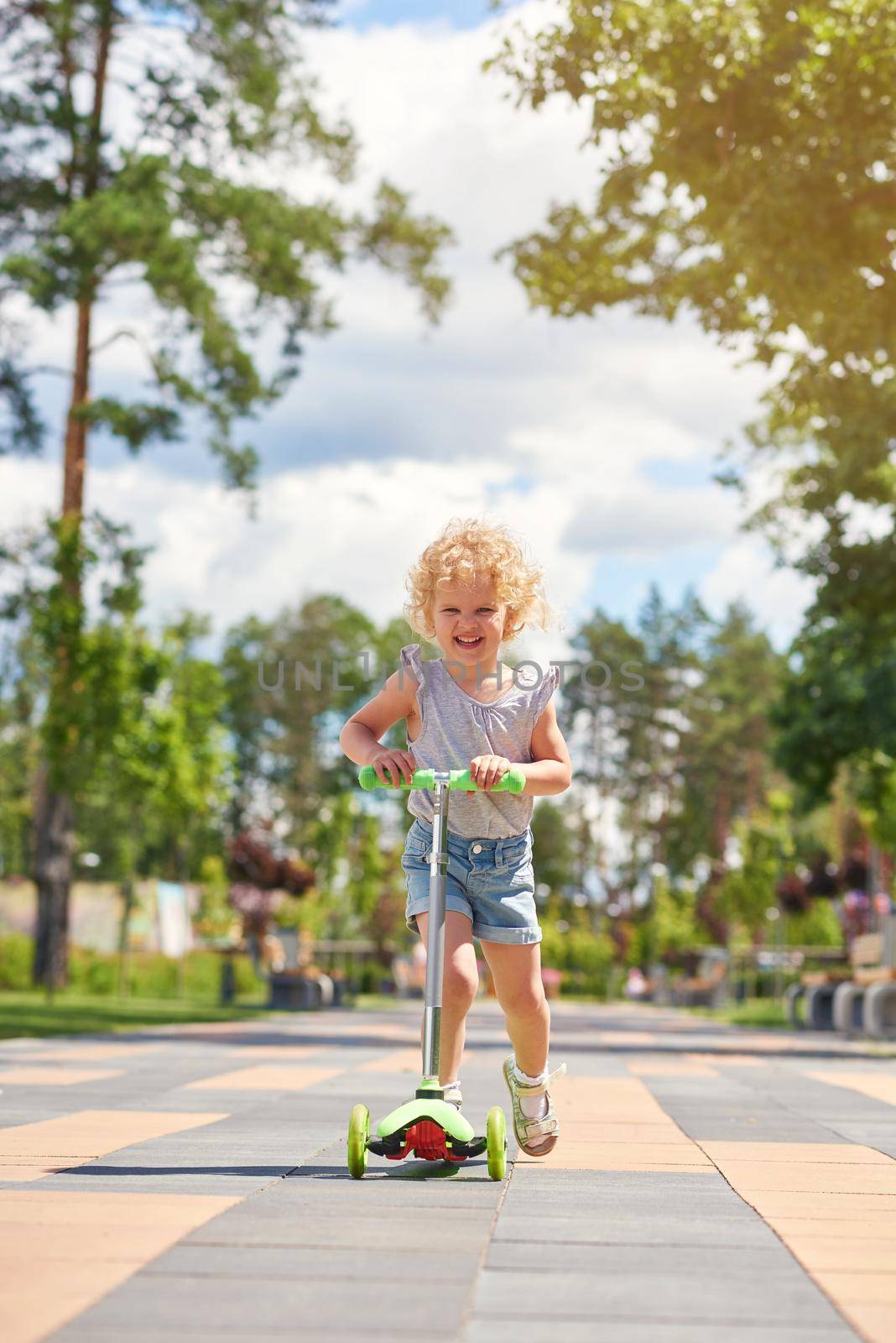 Vertical full length shot of a cute blonde little girl riding scooter at the park kids children happiness emotions activity concept.