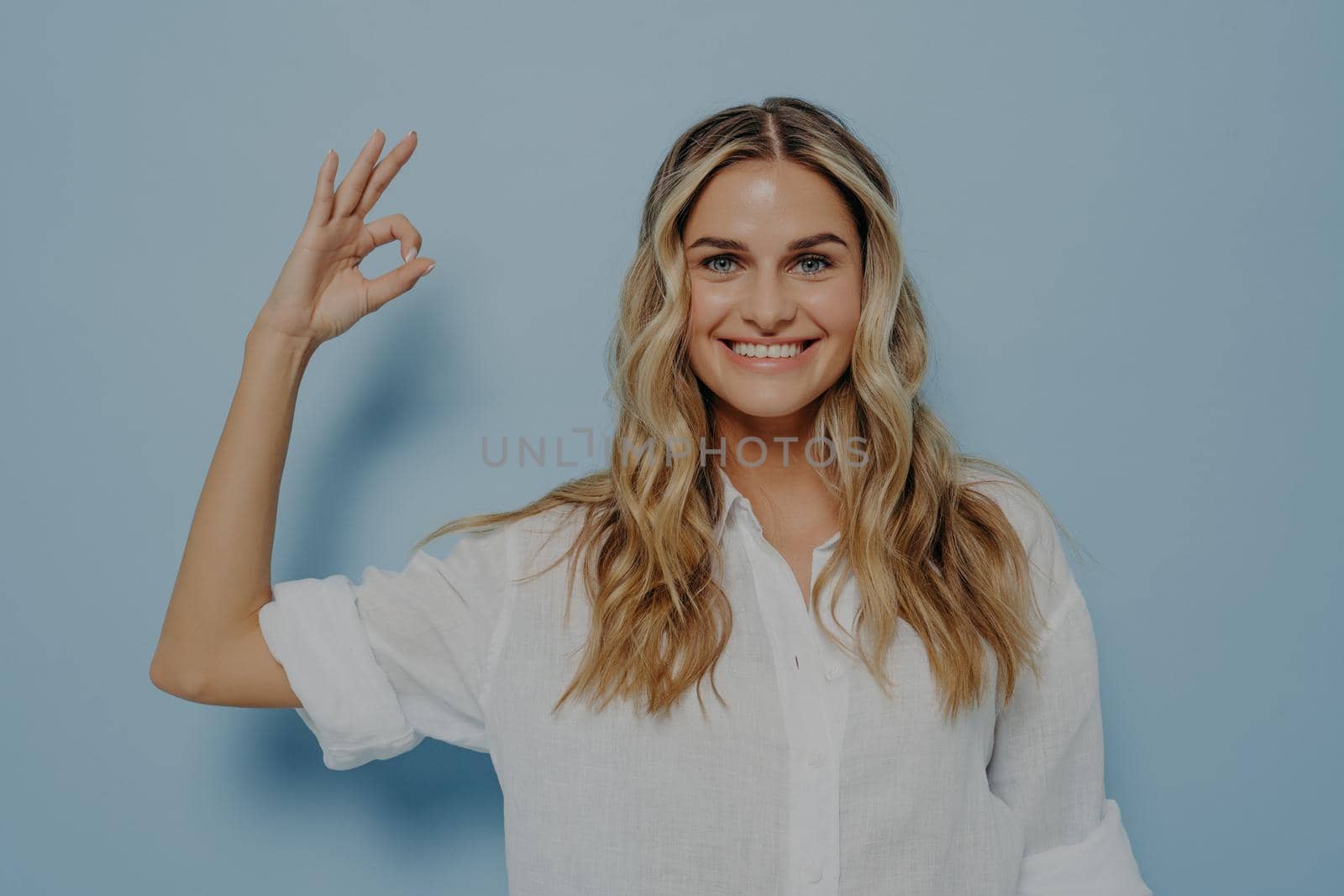 Happy smiling blonde woman showing ok gesture with her hand while smiling, looking forward to having fun and relaxing, standing next to light blue wall. Body language concept