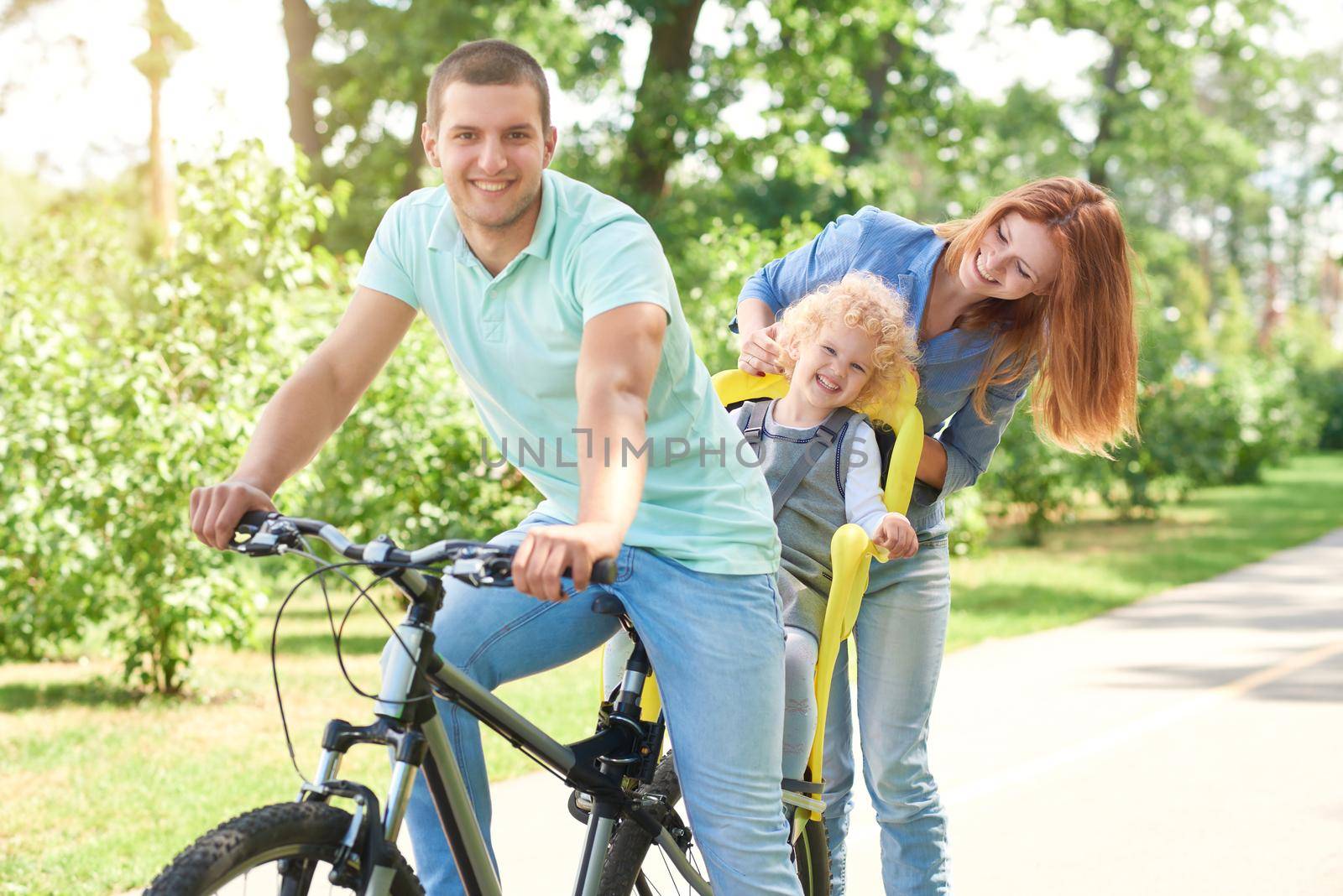 Happy young man smiling to the camera while cycling with his child in a baby bike seat and his beautiful wife at the local park on a warm summer day family active lifestyle concept.