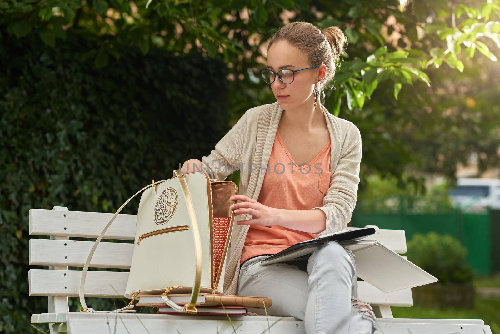 Young girl take something out of her white bag with a golden pattern and a zipper on white wooden bench park in the green city park. She wears blue jeans, cardigan, eyeglasses.