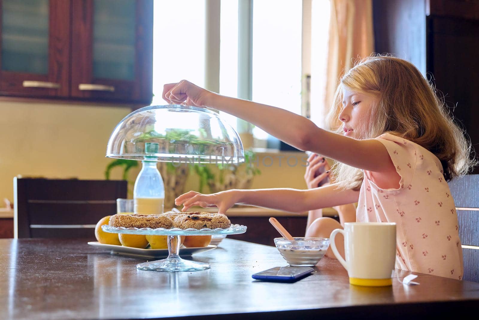 Family, children, two girls, sisters eating at home. Teenagers having breakfast in kitchen, milk fruit snacks on table, older girl with smartphone. Adolescence, home everyday life, real life, lifestyle