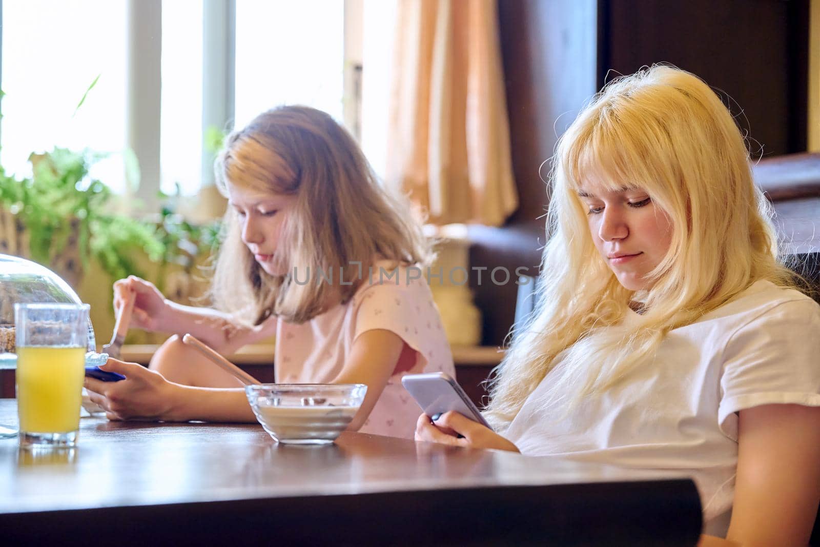 Children two girls sisters eating at home, looking at smartphones. Teenagers having breakfast in kitchen, using phones for leisure entertainment education. Technology in life lifestyle, adolescence
