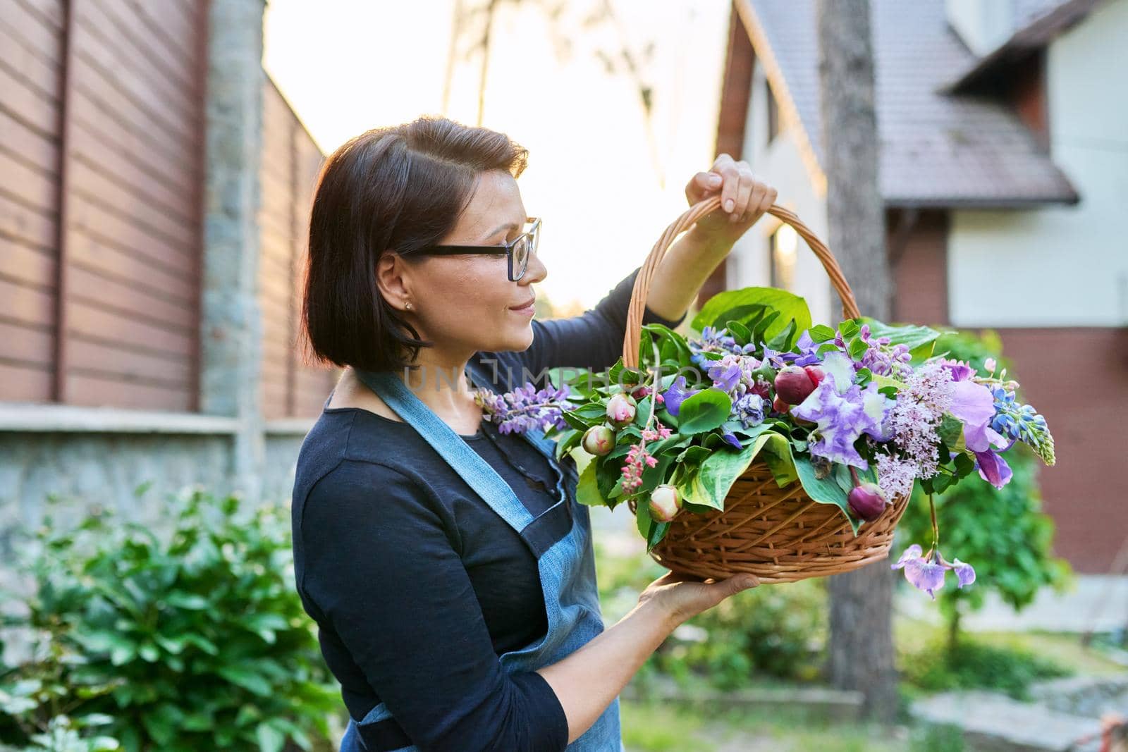 Middle-aged woman gardener florist in an apron with basket of fresh flowers folded in floral arrangement. by VH-studio