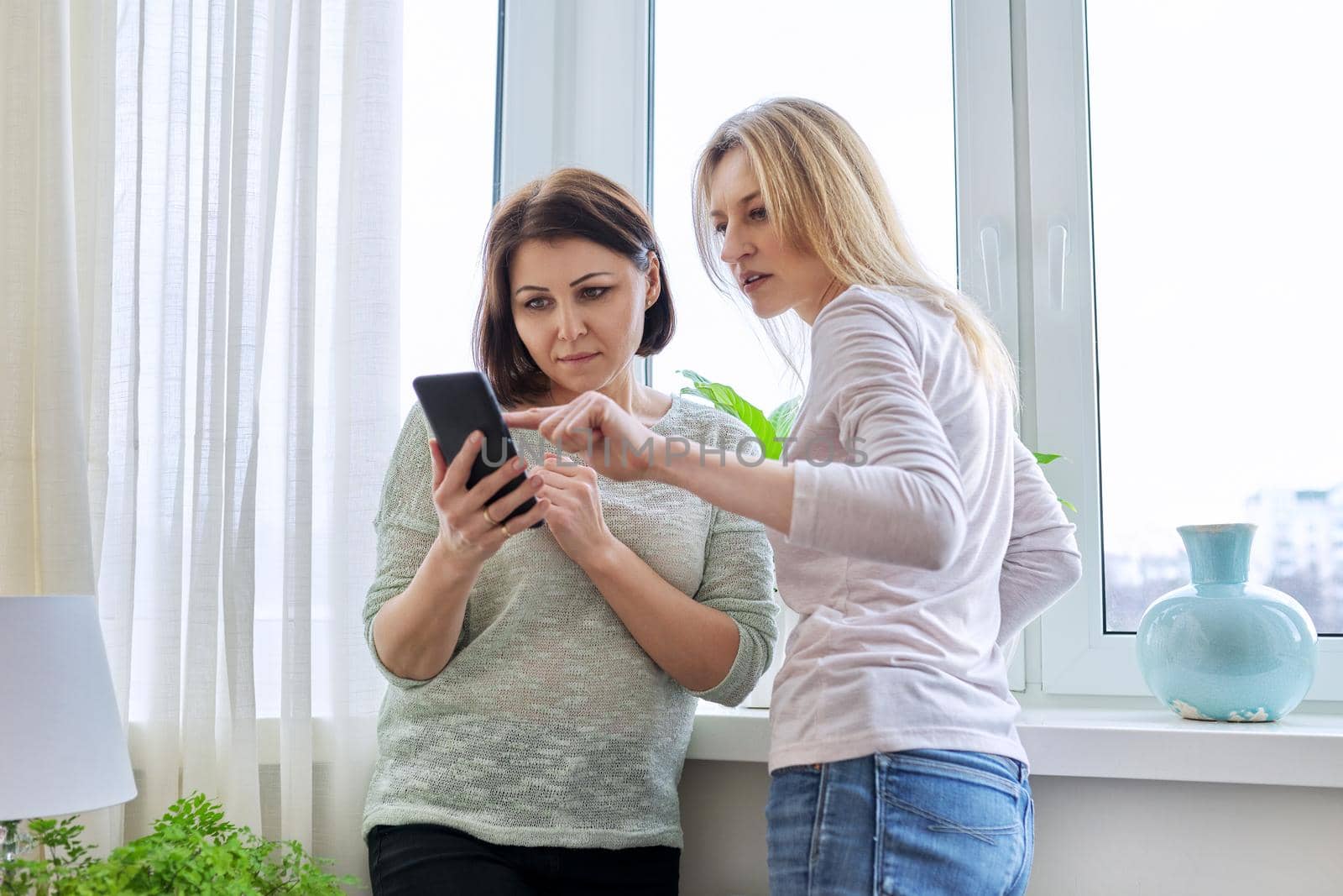 Two middle-aged women friends looking together at smartphone screen. Females at home near window in winter season. Relationship, lifestyle, leisure, friendship, technology, mature people concept