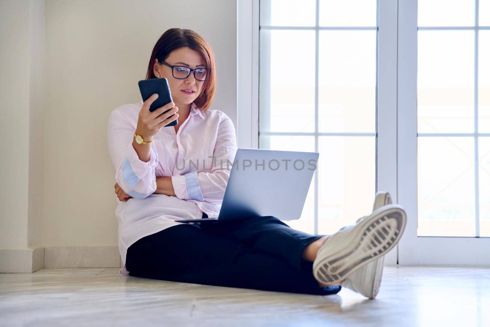Business middle aged woman sitting on the floor with a laptop talking on a mobile phone by VH-studio