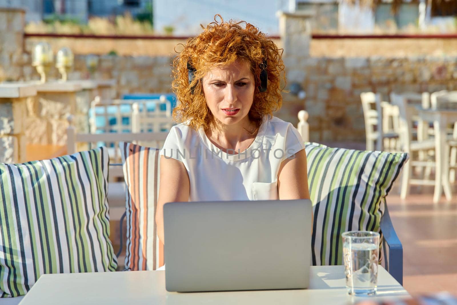 Serious confident woman working with laptop outdoors. Middle aged female with headphones sitting at table in cafe, looking at monitor. Remote work, freelance, online business, video conference, chat