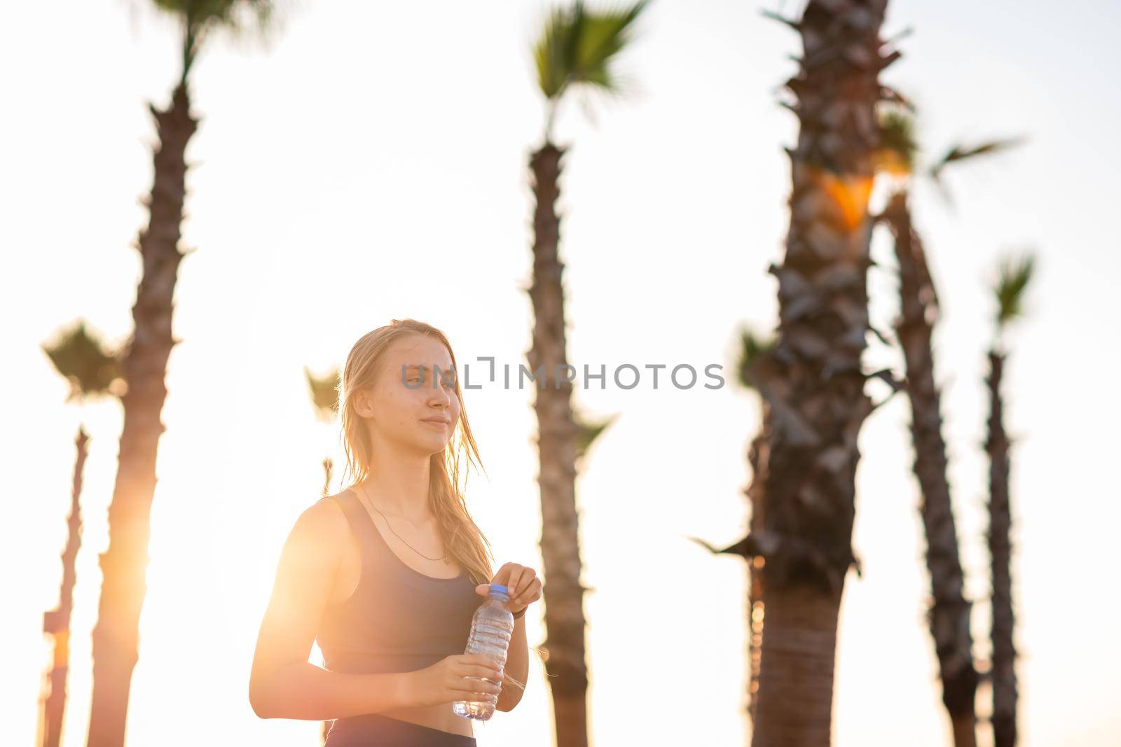 Young girl exercising on seafront line at sunset sunrise. Beautiful girl drinking water while jogging on sea promenade during summer Vacation Meditation Seaside Sea Ocean Holiday Travel