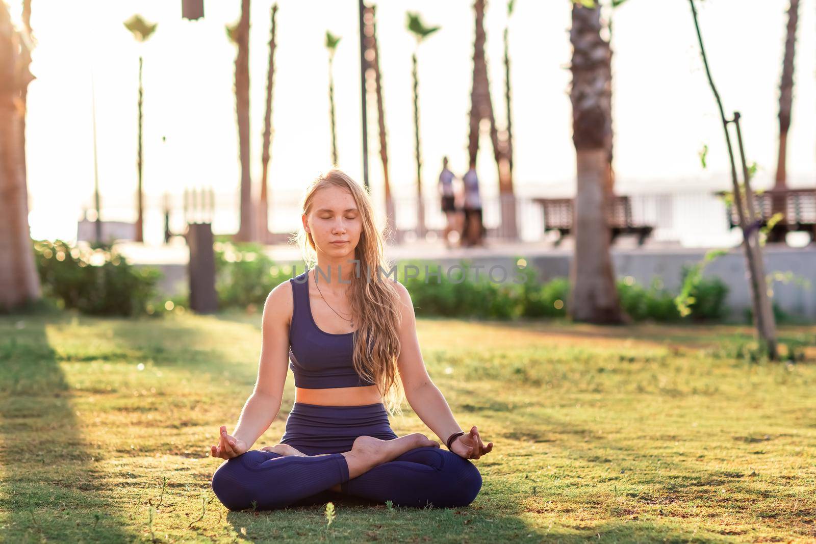 Young girl sitting in Lotus pose in the park near seafront line at sunset sunrise. Beautiful girl practicing Yoga during summer Vacation Meditation Seaside Sea Ocean Holiday Travel