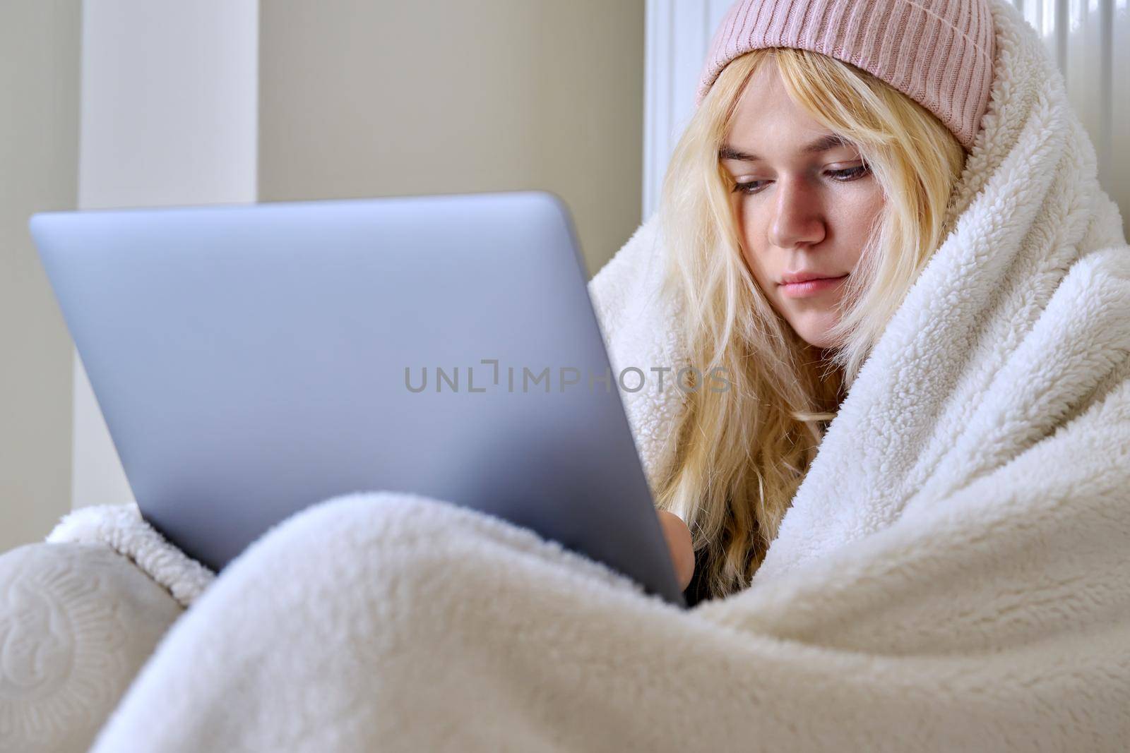 A young female teenager frozen in house in winter cold season, warming up with a warm blanket, hat, central heating radiator, looking at laptop screen, student, having rest