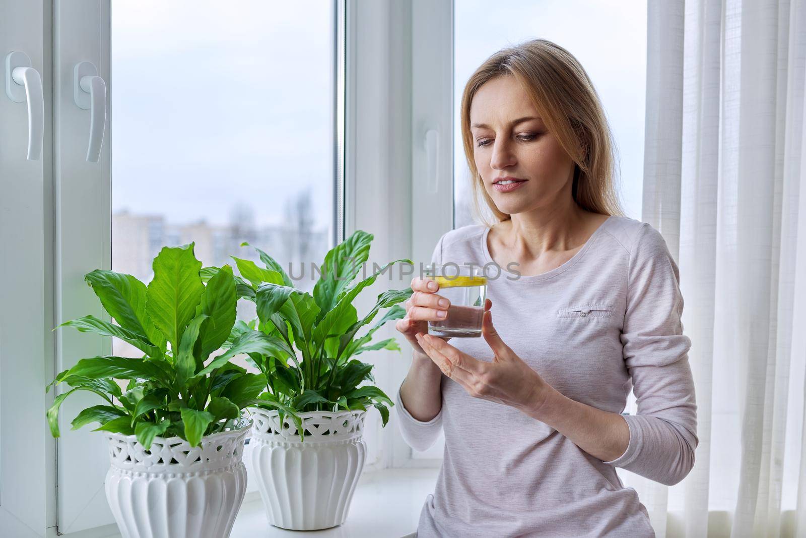 Middle-aged woman drinking lemon water at home near the window, winter autumn season, natural vitamins. Health, nutrition, natural antioxidant, beauty, 40s, mature people, lifestyle concept