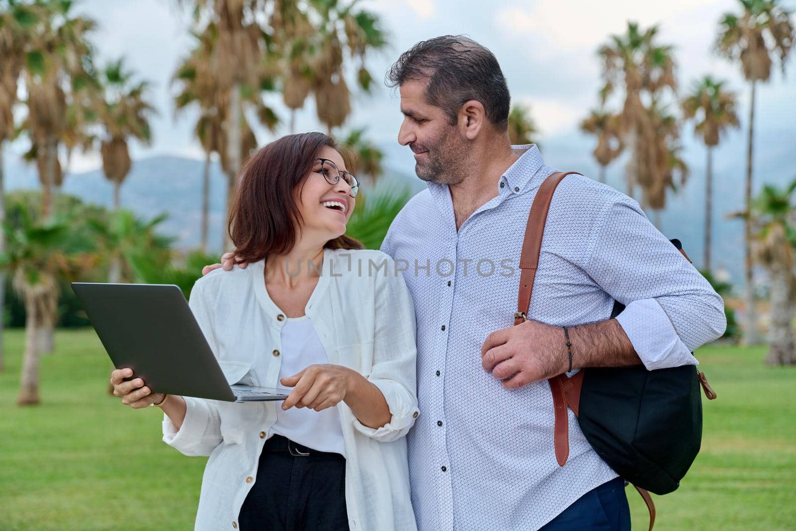 Middle-aged man and woman looking in laptop screen outdoors. Couple of business colleagues talking discussing smiling, tropical park nature background. Business, people, technology, mature age concept