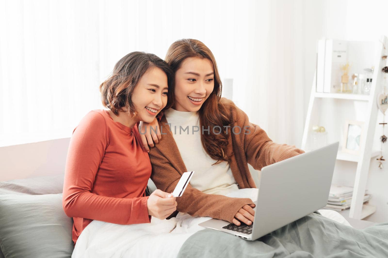 Two friends shopping online with credit card and laptop lying on the bed at home