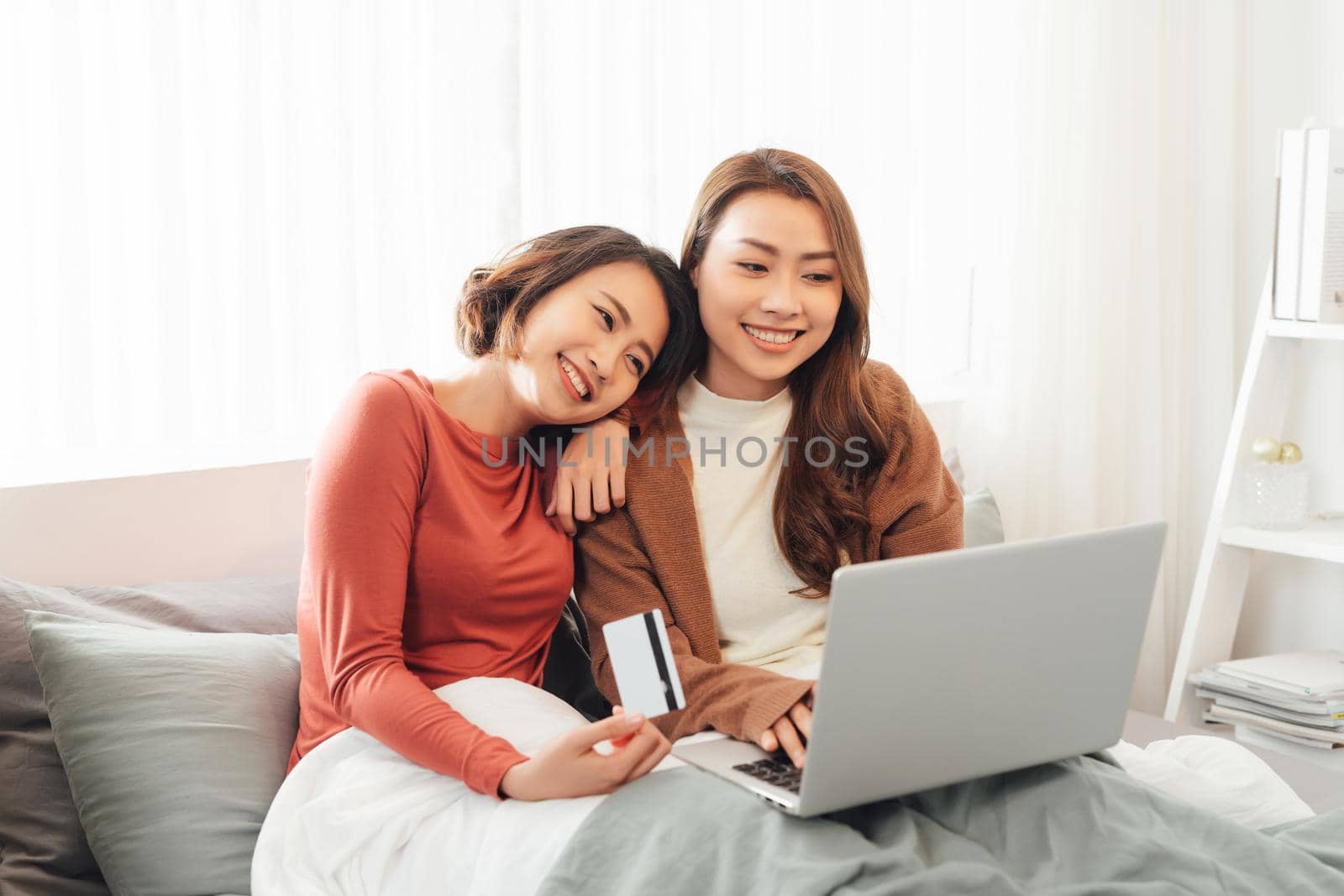 Two beautiful women lying in bed, holding a credit card and a laptop in it