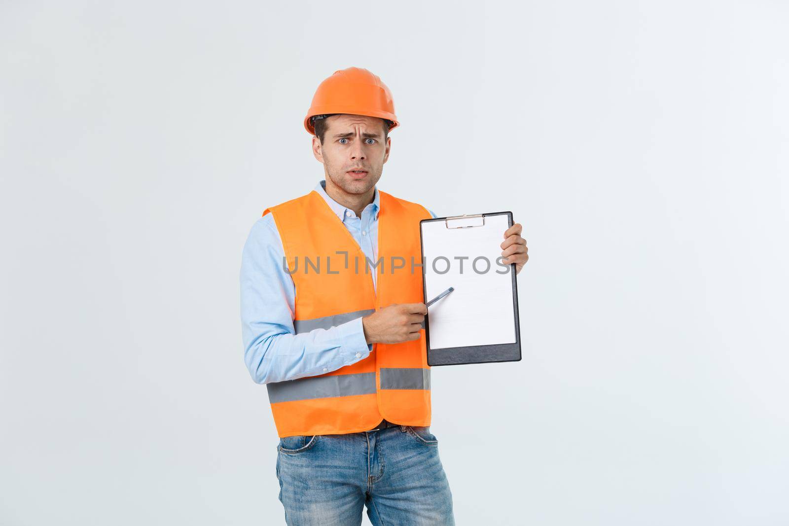Frustrated young engineer with hardhat and reflective vest checking on mistake in document over gray background.