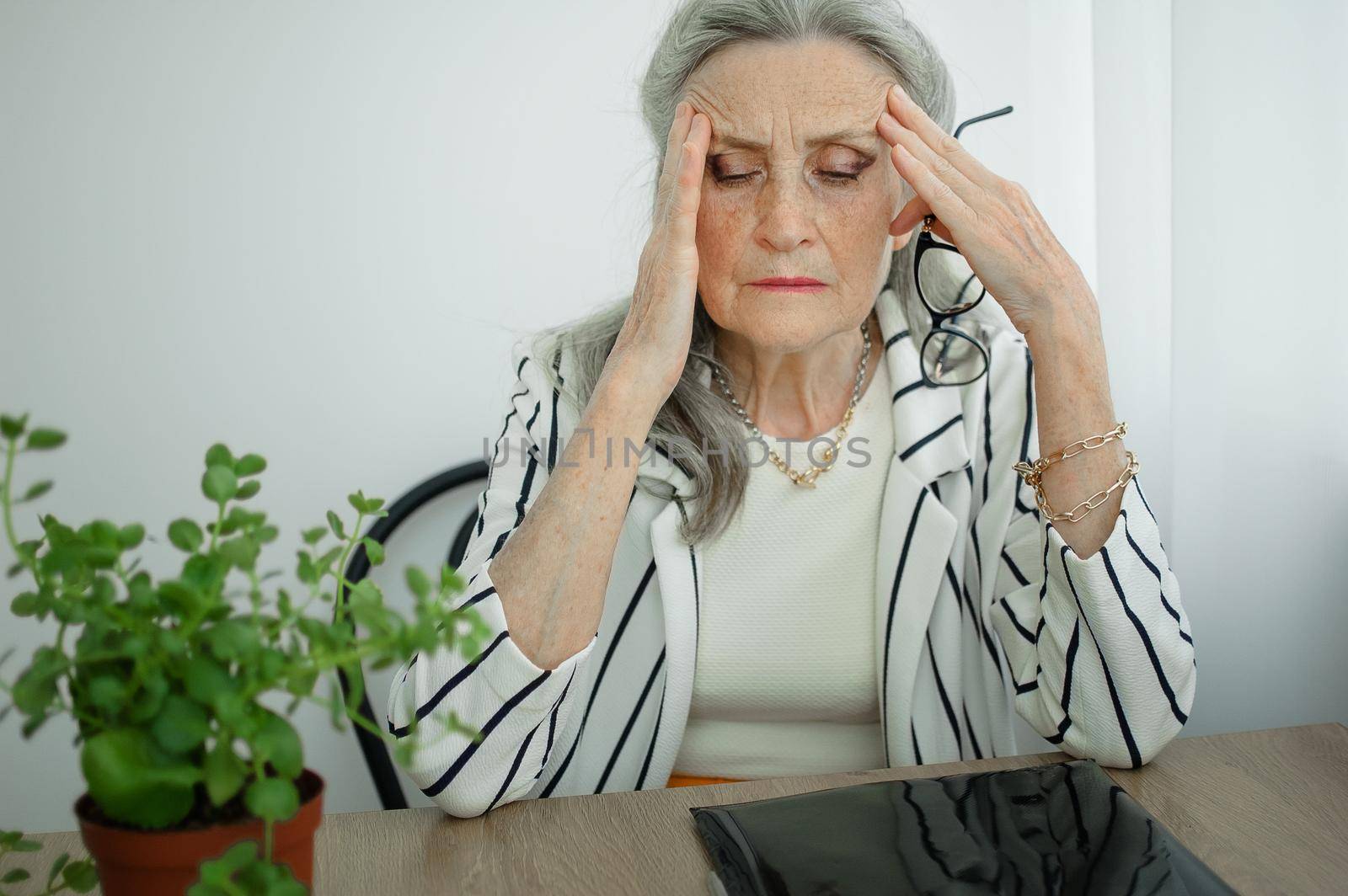 Tired senior grey haired businesswoman in striped jacket with eyeglasses is working in her office sitting at the desk and feeling bad due to menopause, menopause relief concept by balinska_lv