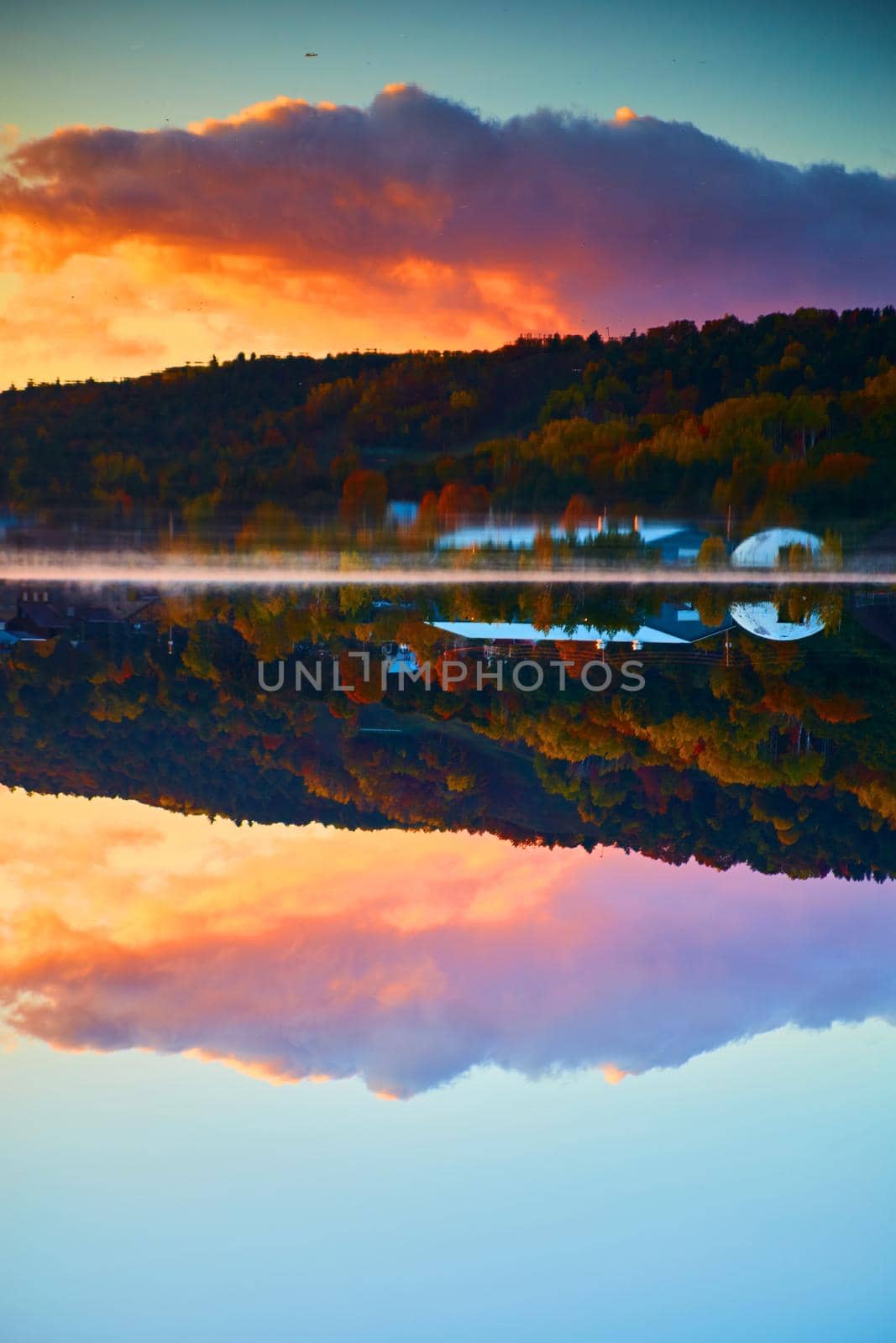 Image of Vertical of a blue sky sunrise with purple pink and orange clouds with a mirror rection of water and a house or barn with fall trees