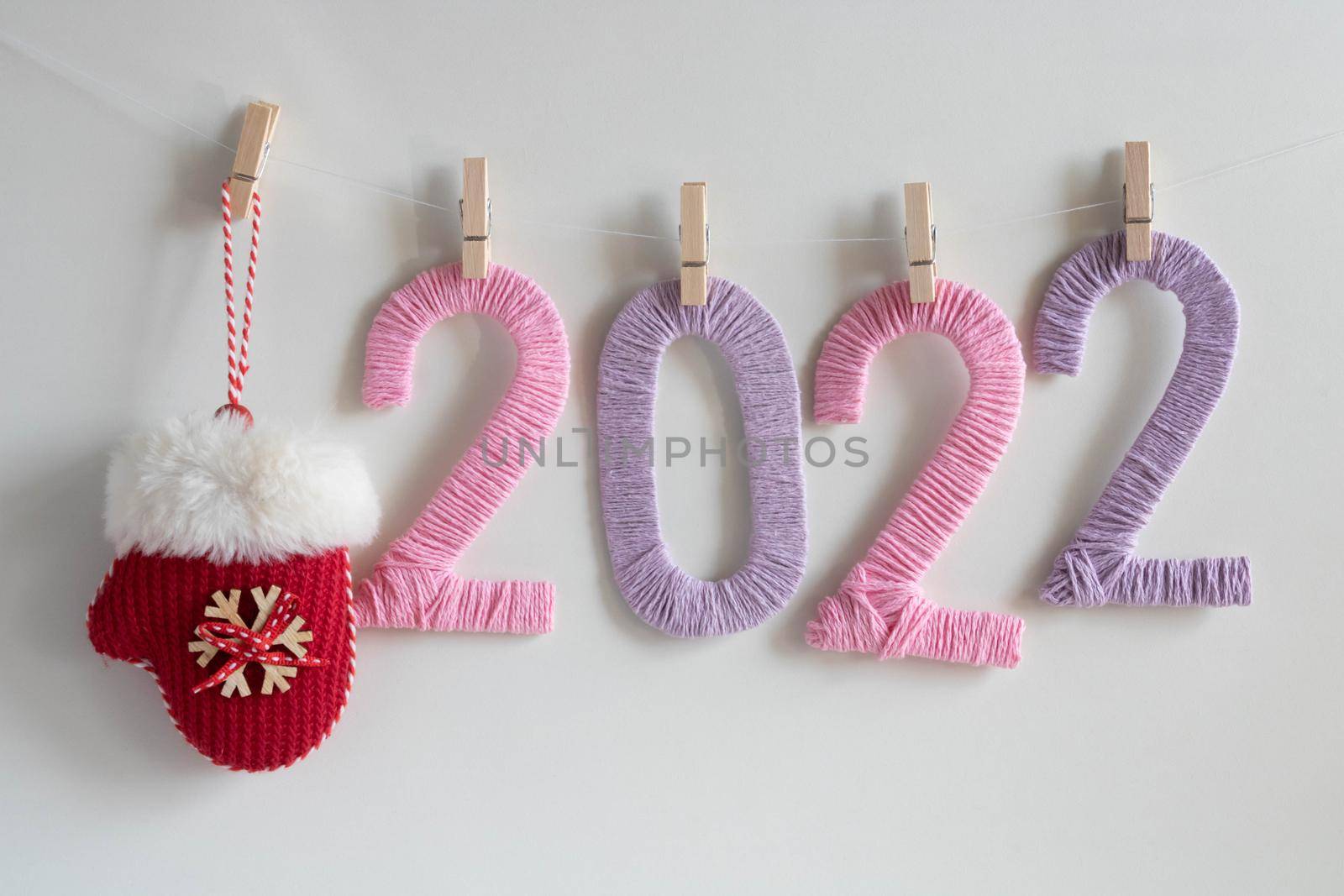 Knitted figures of 2022, made of pink and lilac threads, hang on clothespins on a white background with a red mitten. The concept of the New Year.