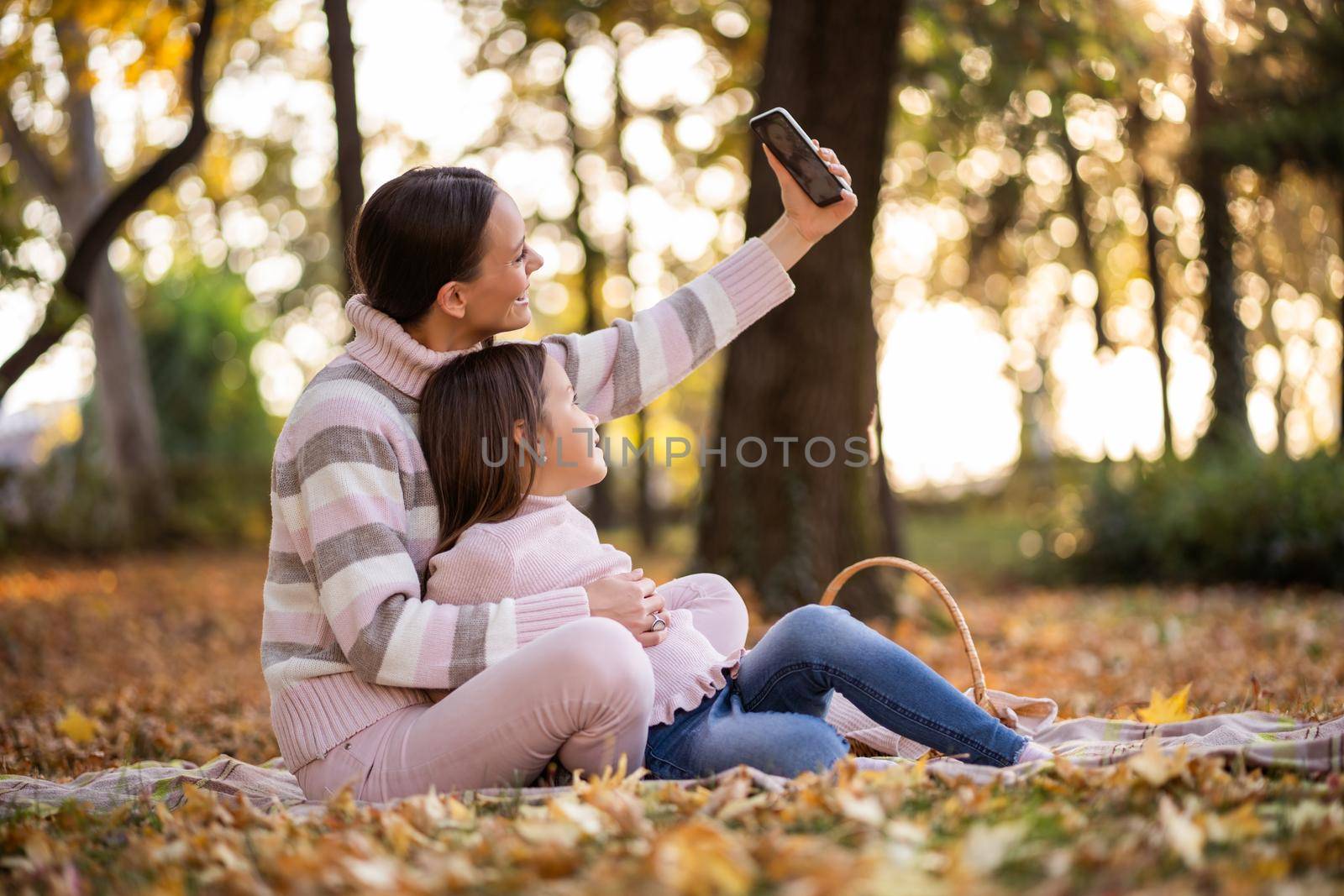 Mother and daughter using smartphone in autumn in park. They are taking selfie.