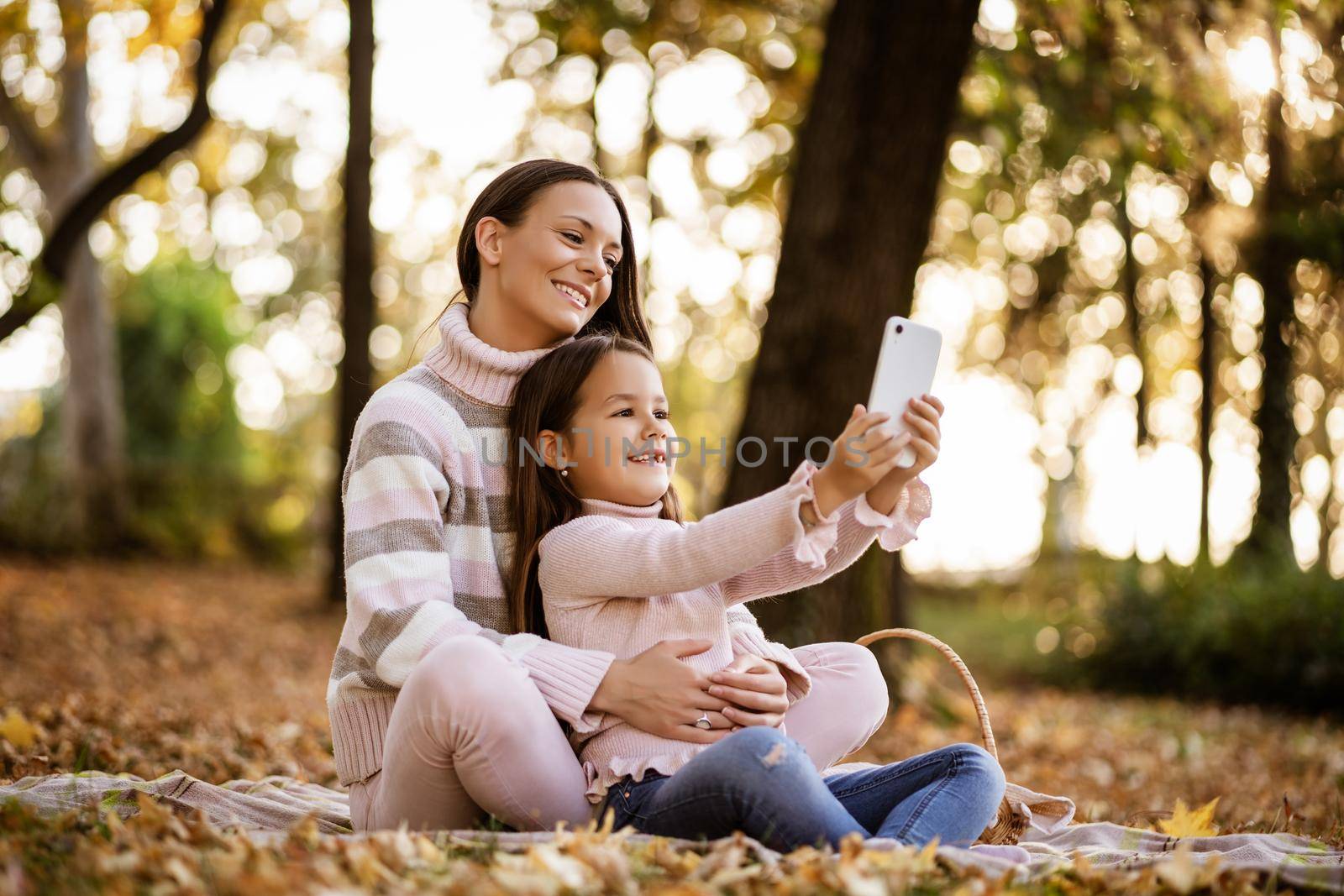 Mother and daughter using smartphone in autumn in park. They are taking selfie.