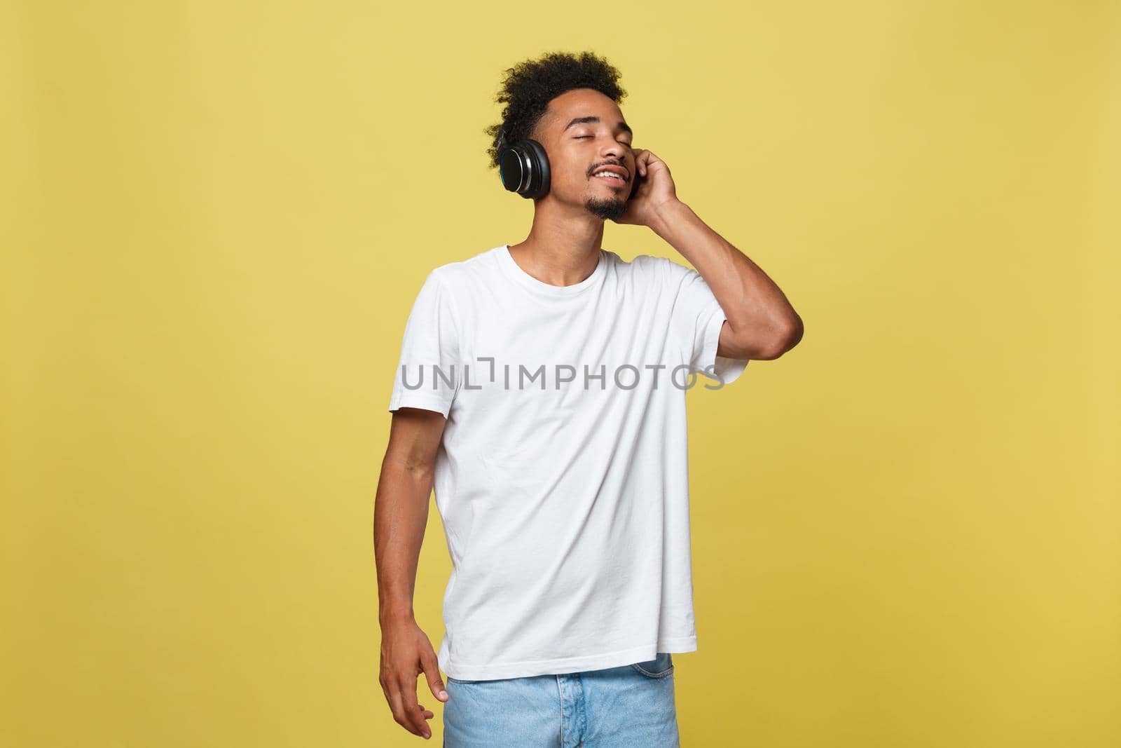 Close up portrait of dark skinned man with closed eyes, has headphones, listens to favourite music. Teenager enjoys audio track isolated on yellow background